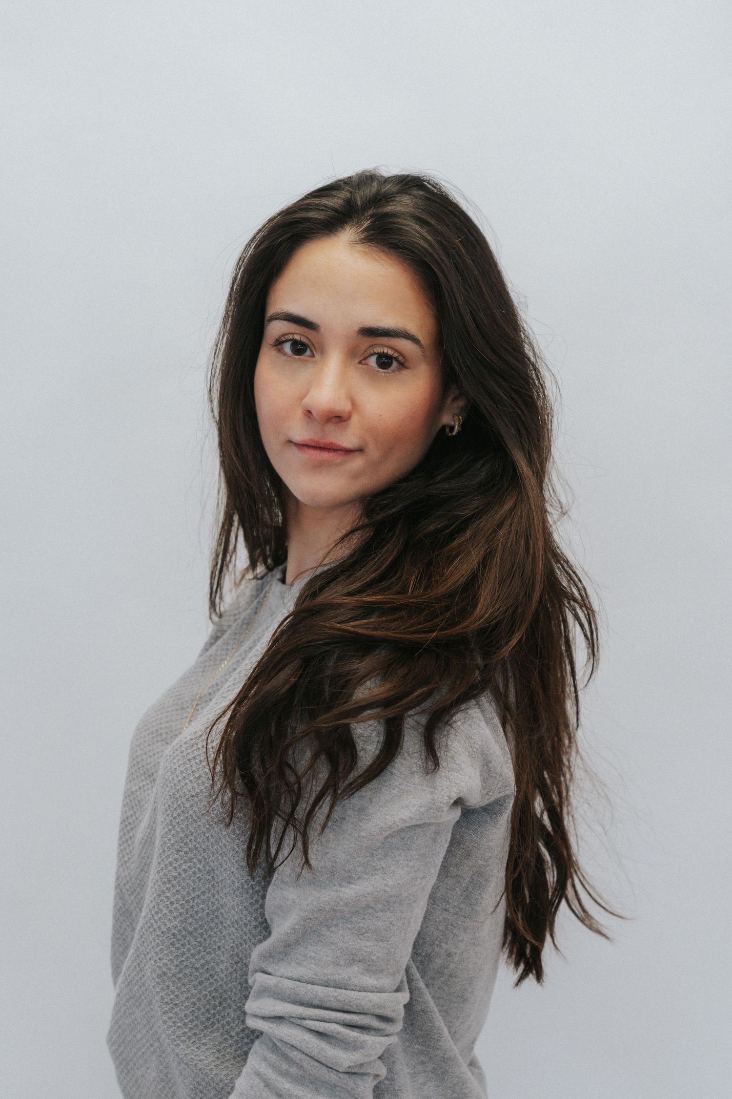 Bruna Mascarenhas Harris - Photographer wearing a gray sweater poses against a white background with long dark hair flowing to one side.