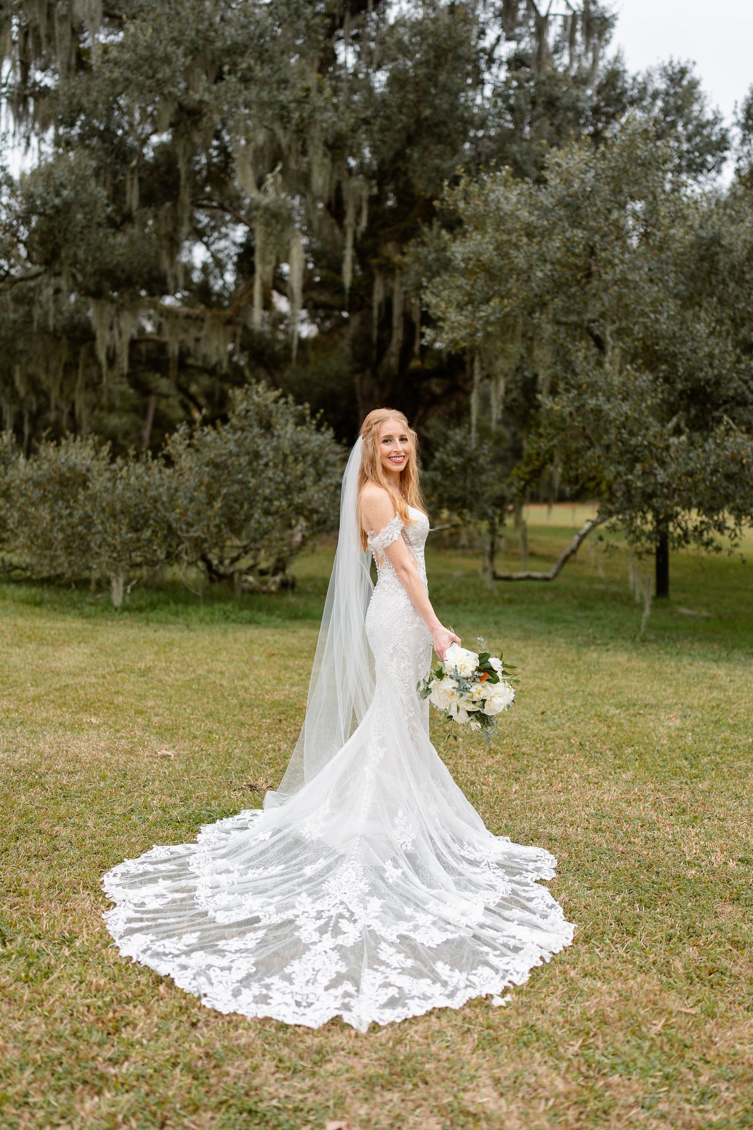 Bride poses with bouquet in elegant lace wedding gown with detailed train in garden setting.