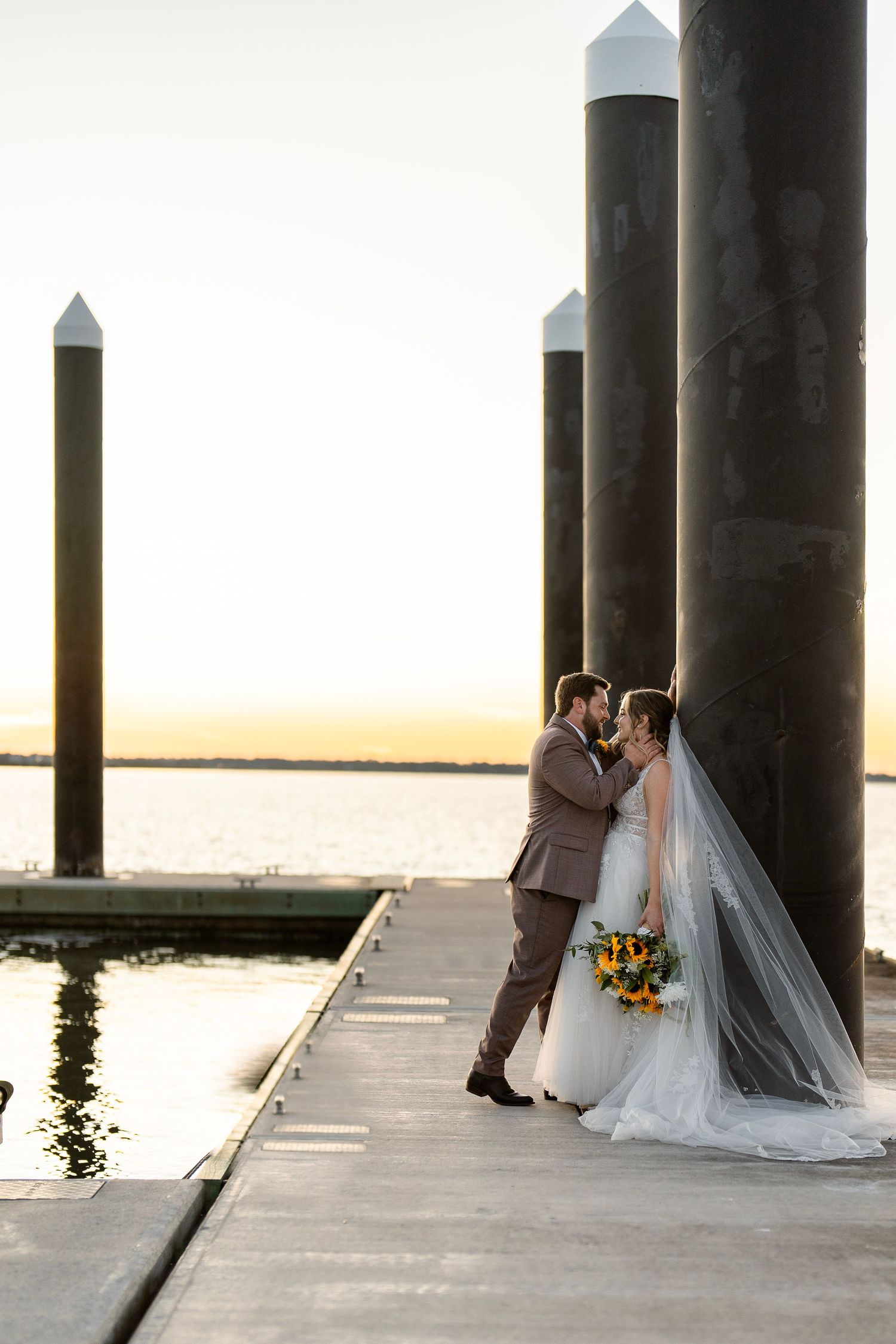 Newlyweds share intimate moment on dock with waterfront sunset and sunflower bouquet.