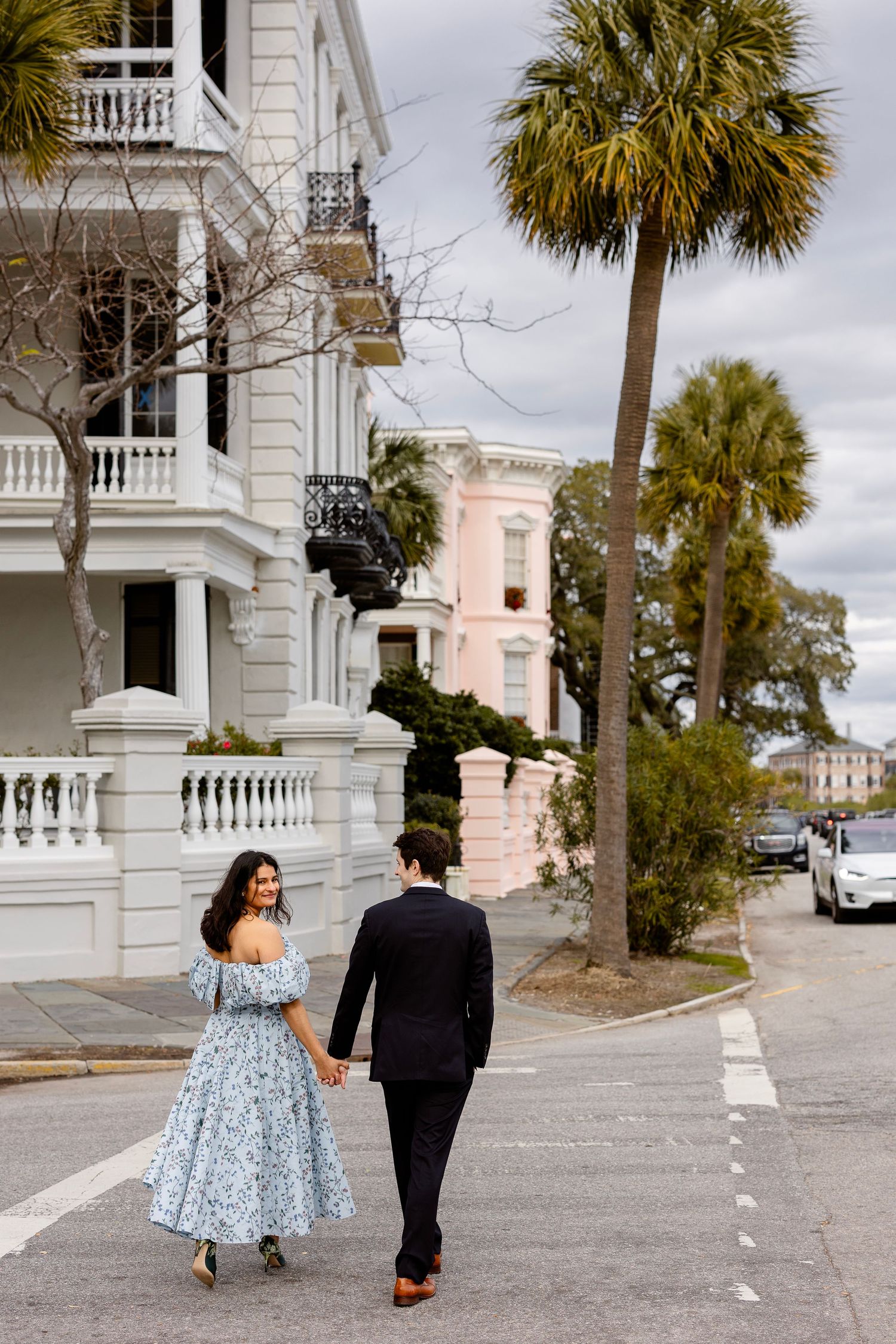 A couple strolls down a historic Charleston street with palm trees and colorful architecture.