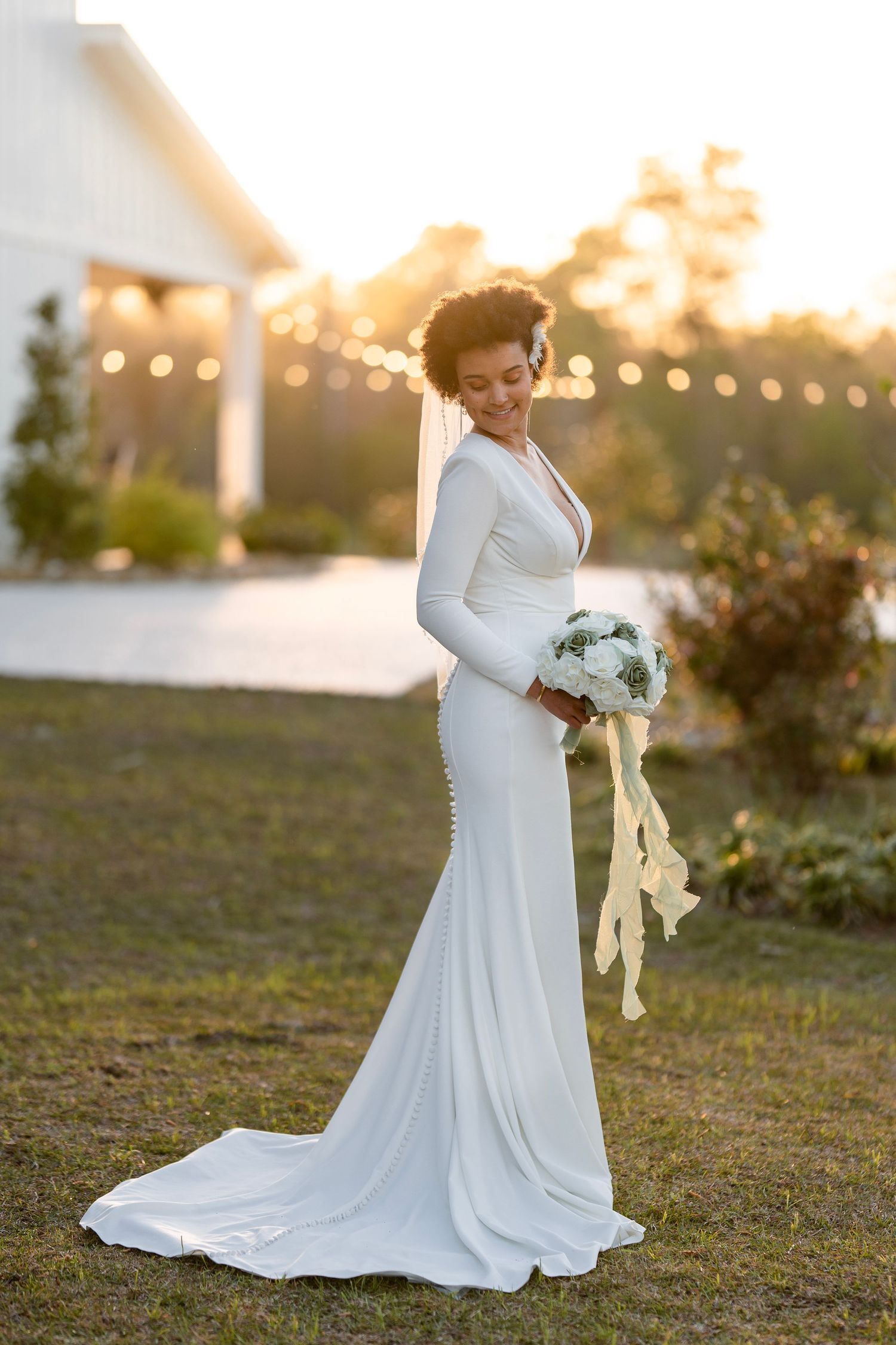 Bride in sleek white long-sleeve gown poses at golden hour with floral bouquet.