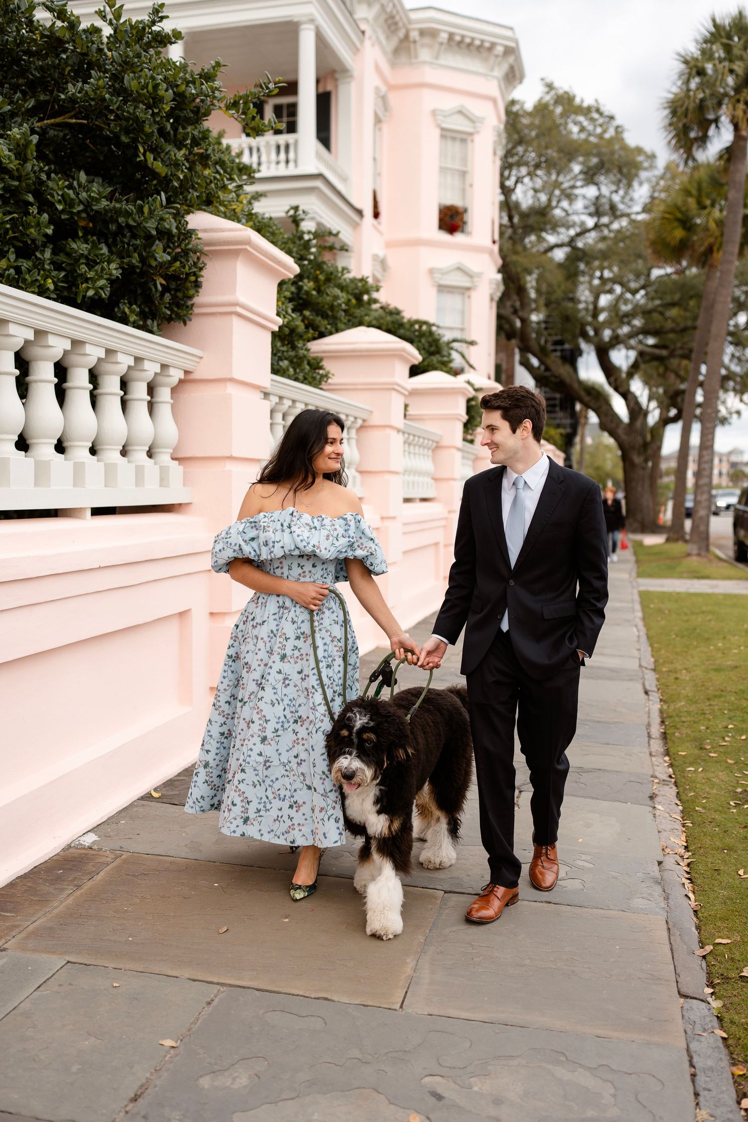 A couple walks their fluffy dog along a pink antebellum mansion's sidewalk in Charleston.