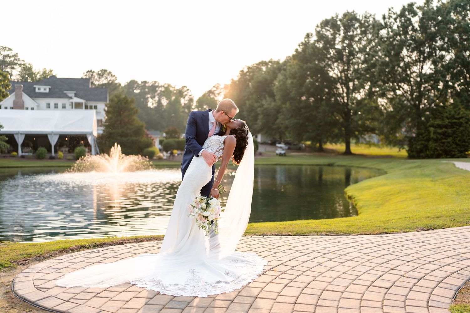 Sunset wedding portrait on brick patio beside lake with white house in background.
