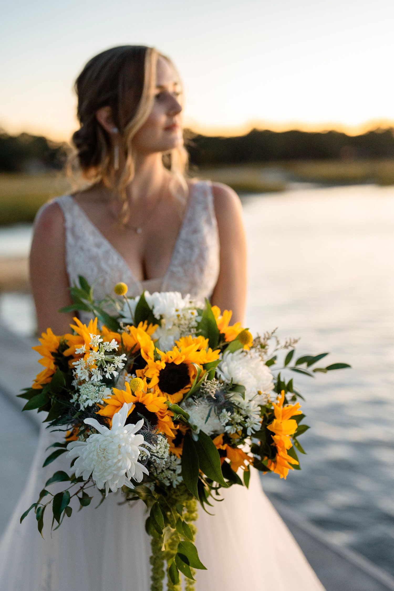 Wedding bouquet with bright sunflowers and white flowers held against sunset lake backdrop.