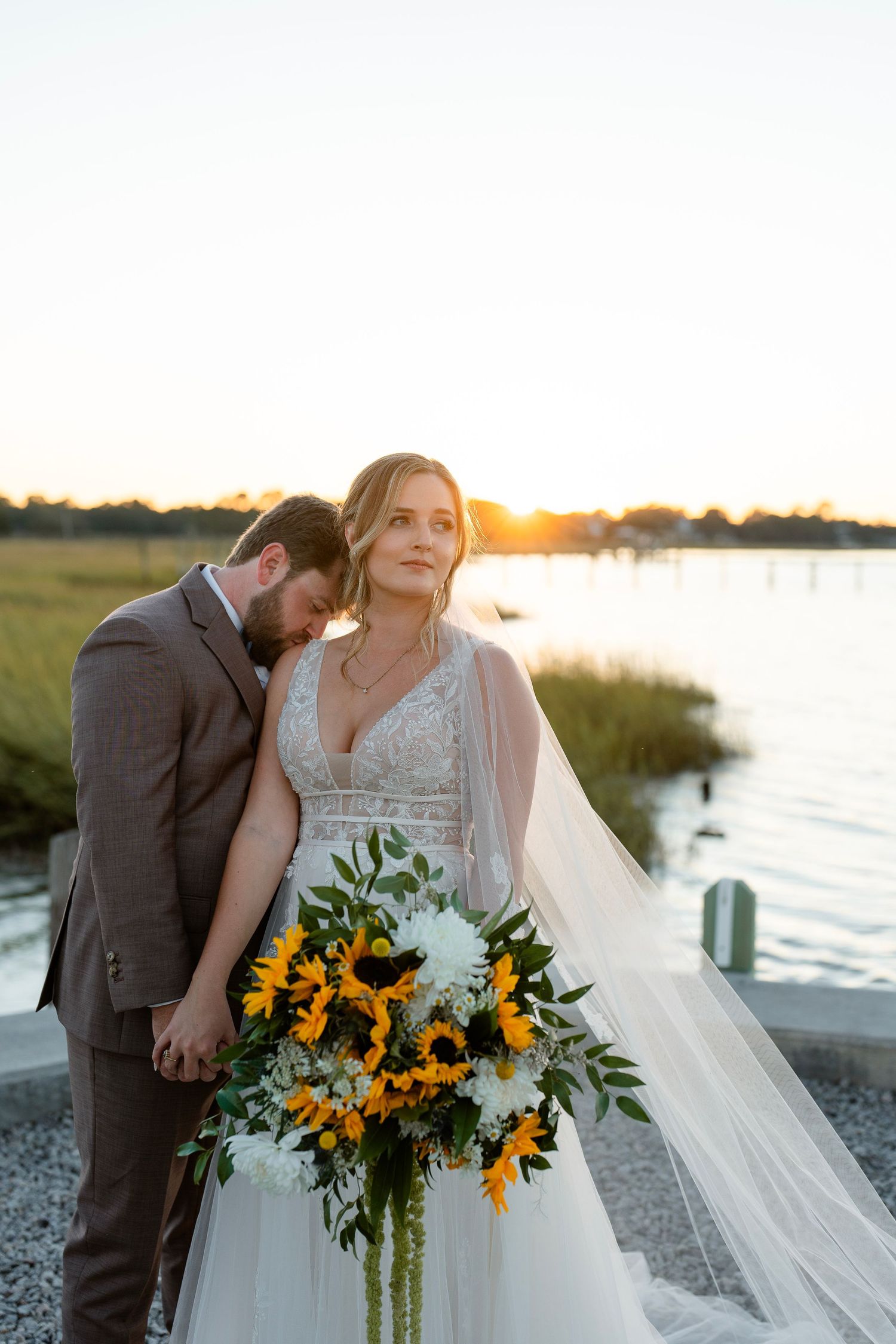 Wedding couple embracing at sunset by waterfront with sunflower bouquet.