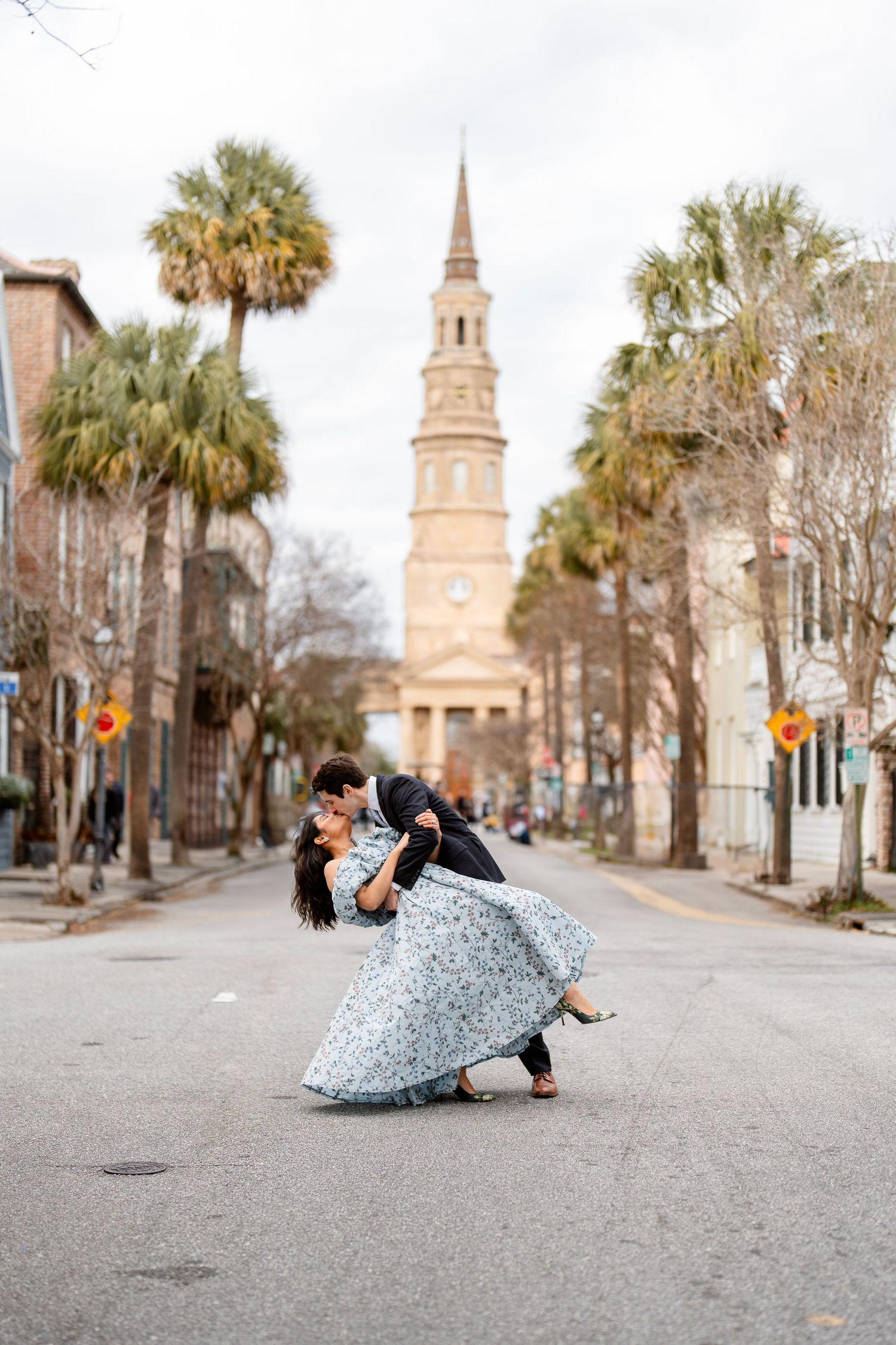 Couple dips while dancing in front of historic Charleston church with palm trees lining the street.