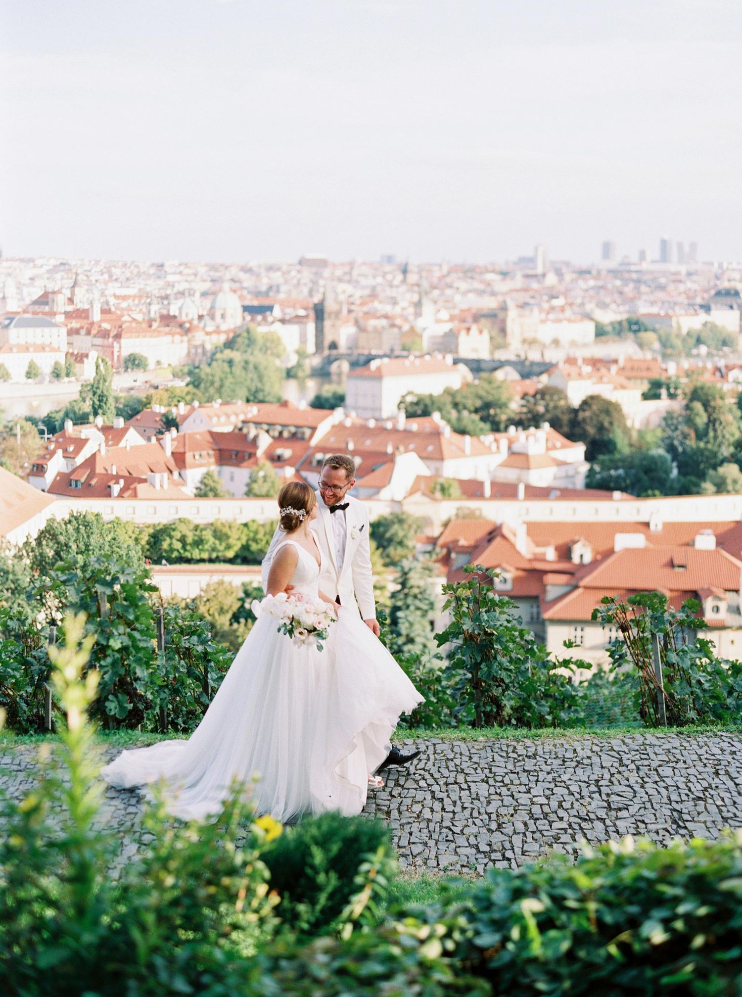 Bride and groom overlook Prague city skyline from lush garden terrace in elegant wedding attire.
