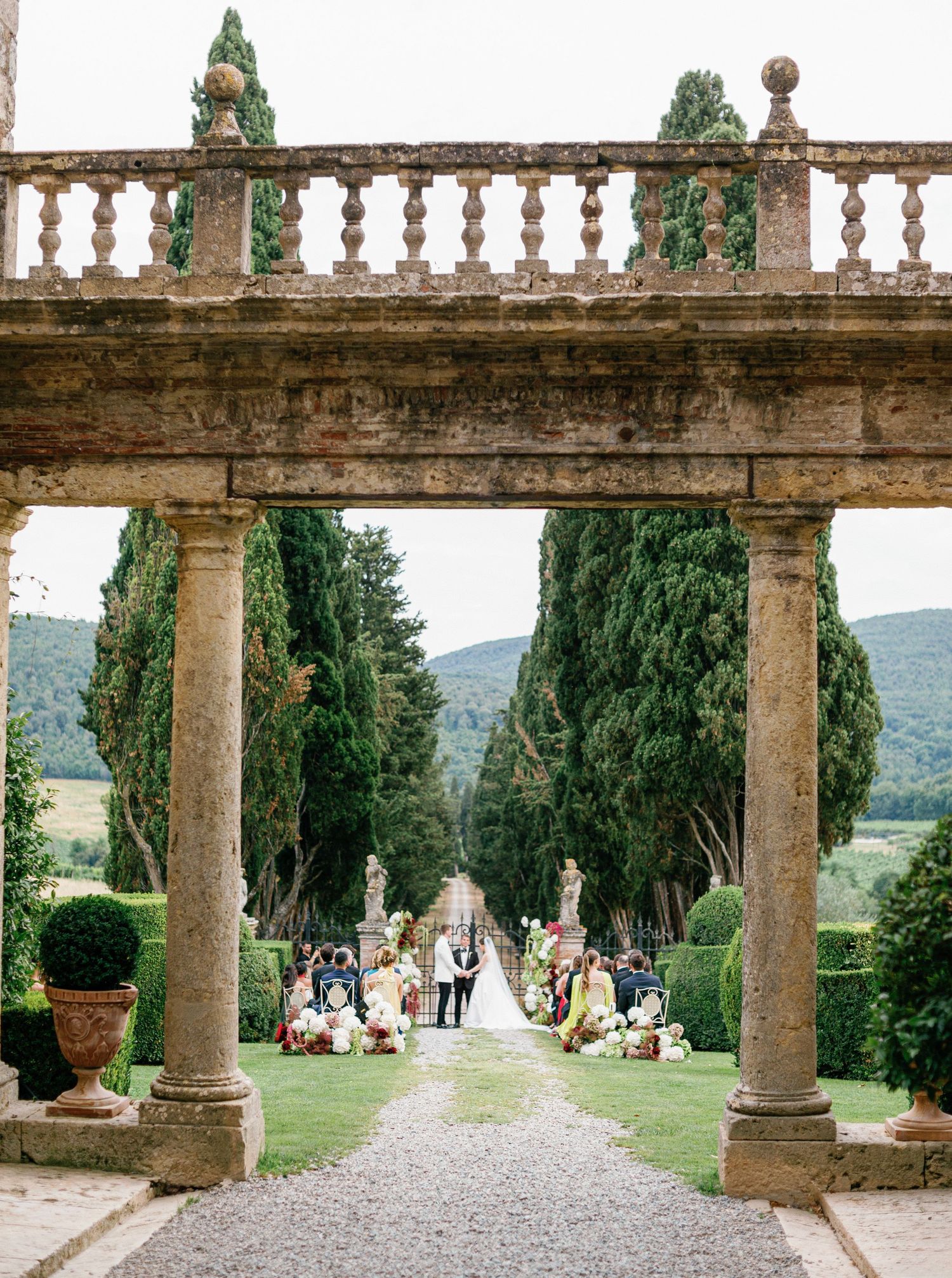 Stone columns line a scenic garden pathway at a grand Italian villa Borgo Stomennano with cypress trees and mountains in the distance.
