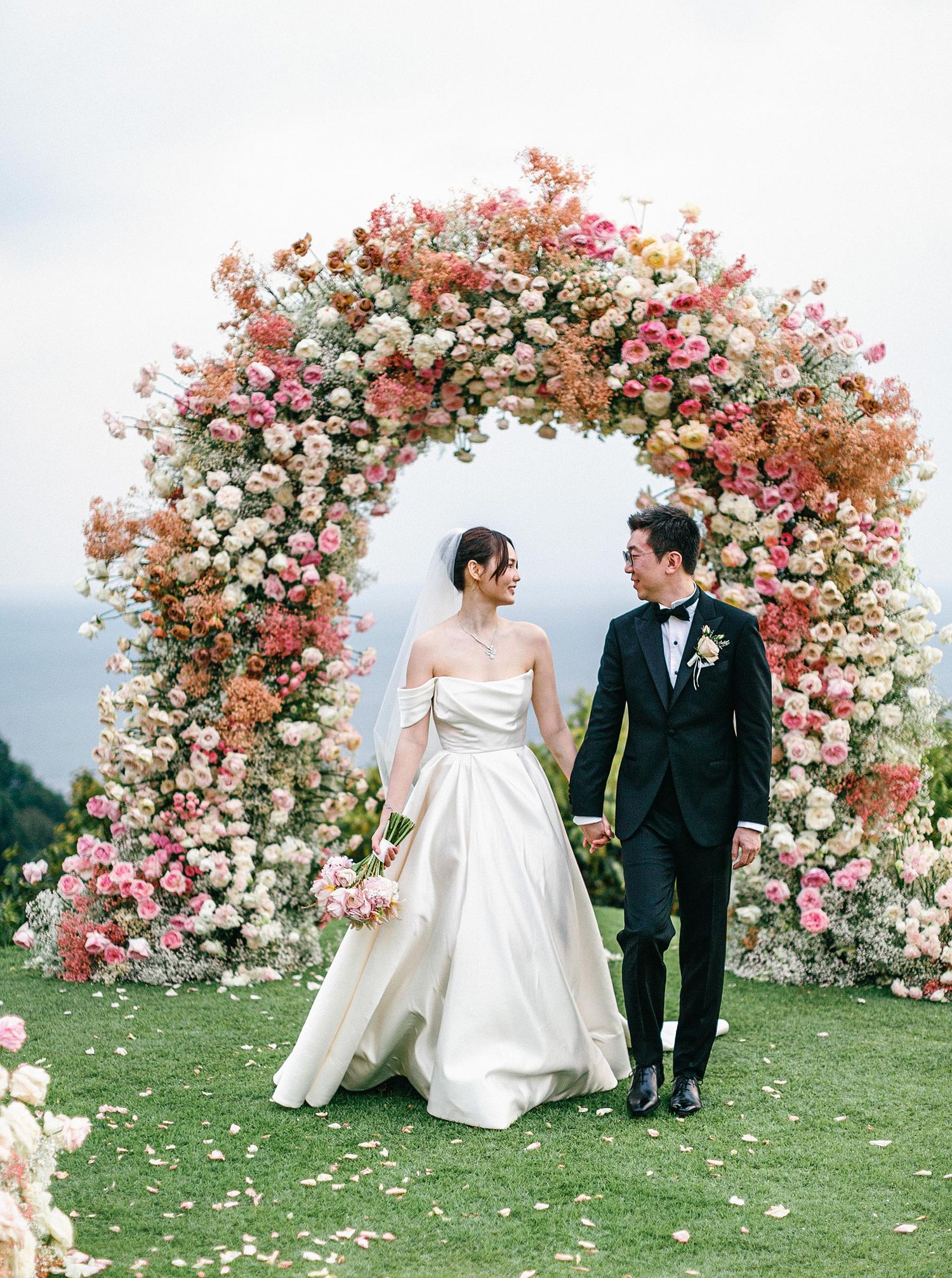 Elegant wedding couple at Paresa, Phuket, Thailand, walking under a colorful floral arch with ocean views.