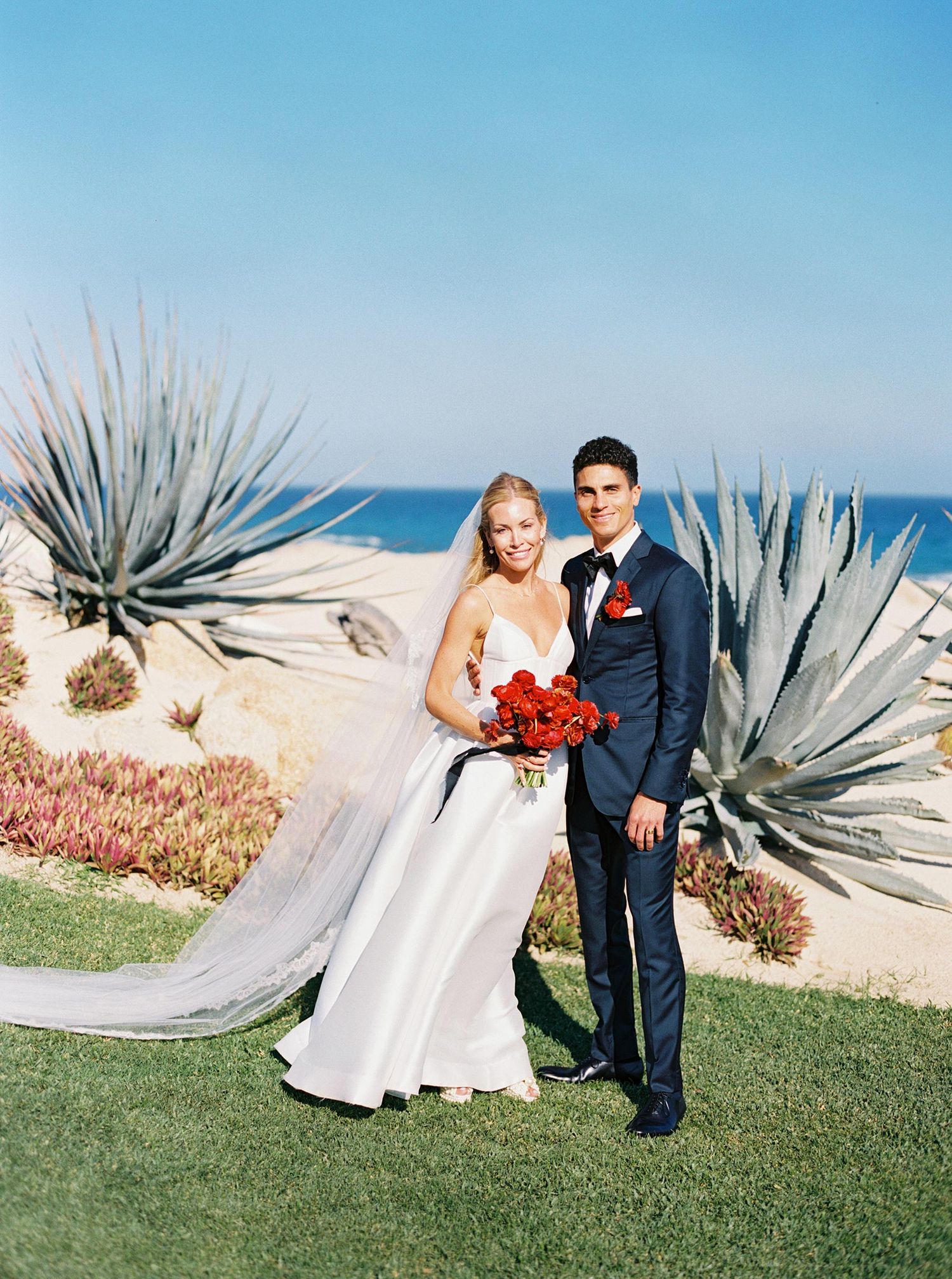 Bride and groom pose together outdoors near agave plants with ocean views, she holds red roses bouquet.