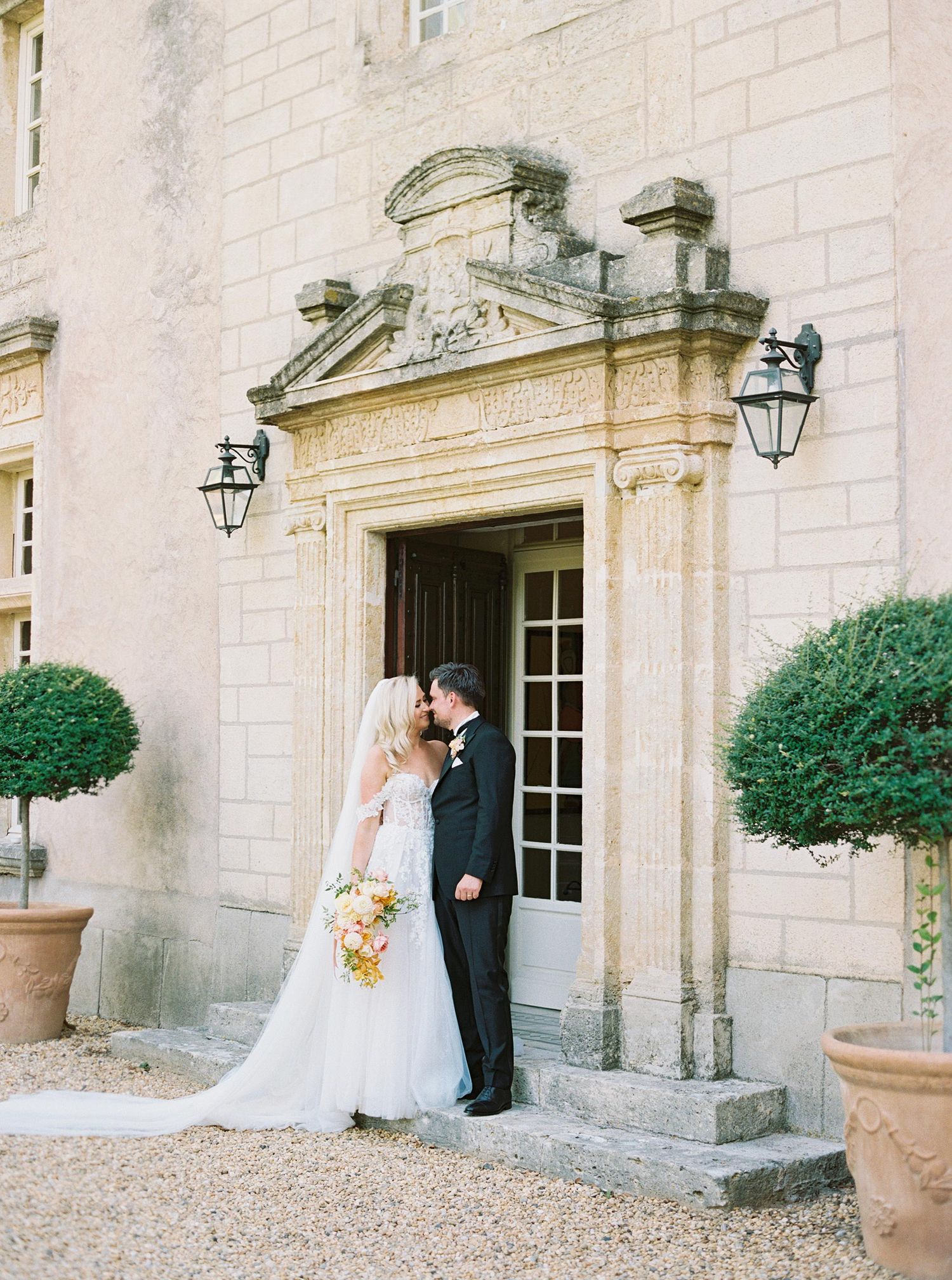 An elegant couple shares an intimate moment by a grand limestone entrance with ornate architectural details at a French chateau.