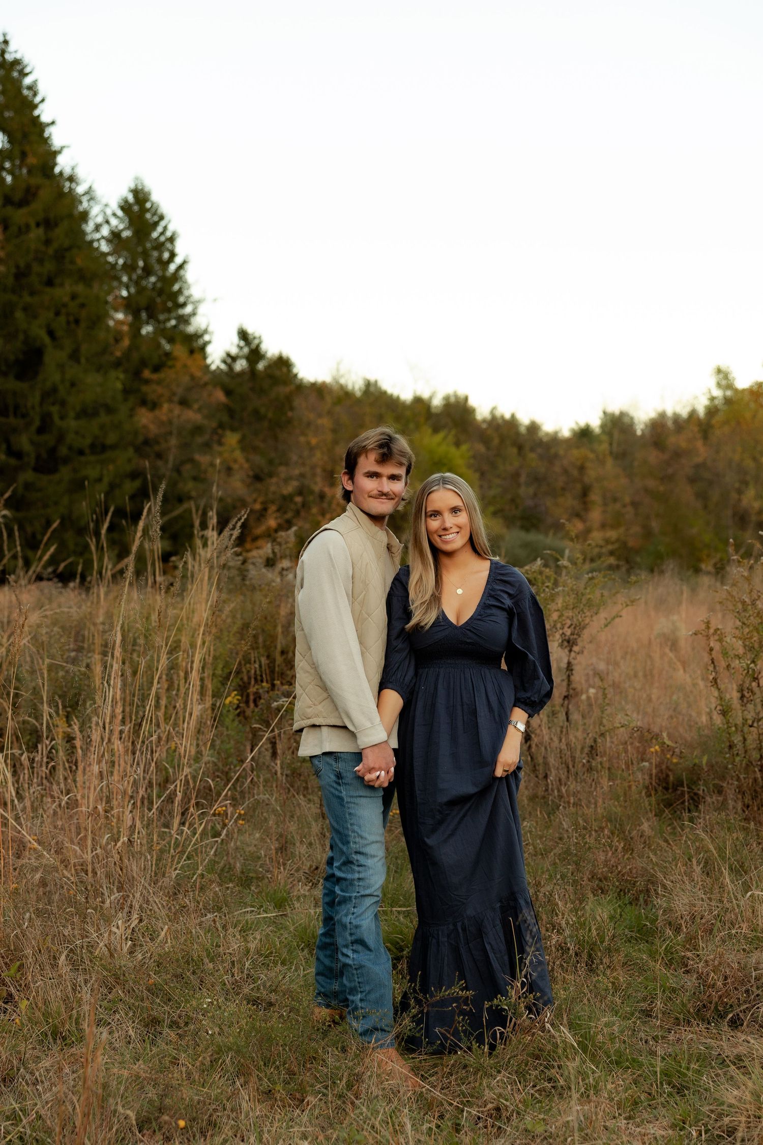 A couple shares a romantic moment together in a rustic field with tall grass and evergreen trees at sunset.