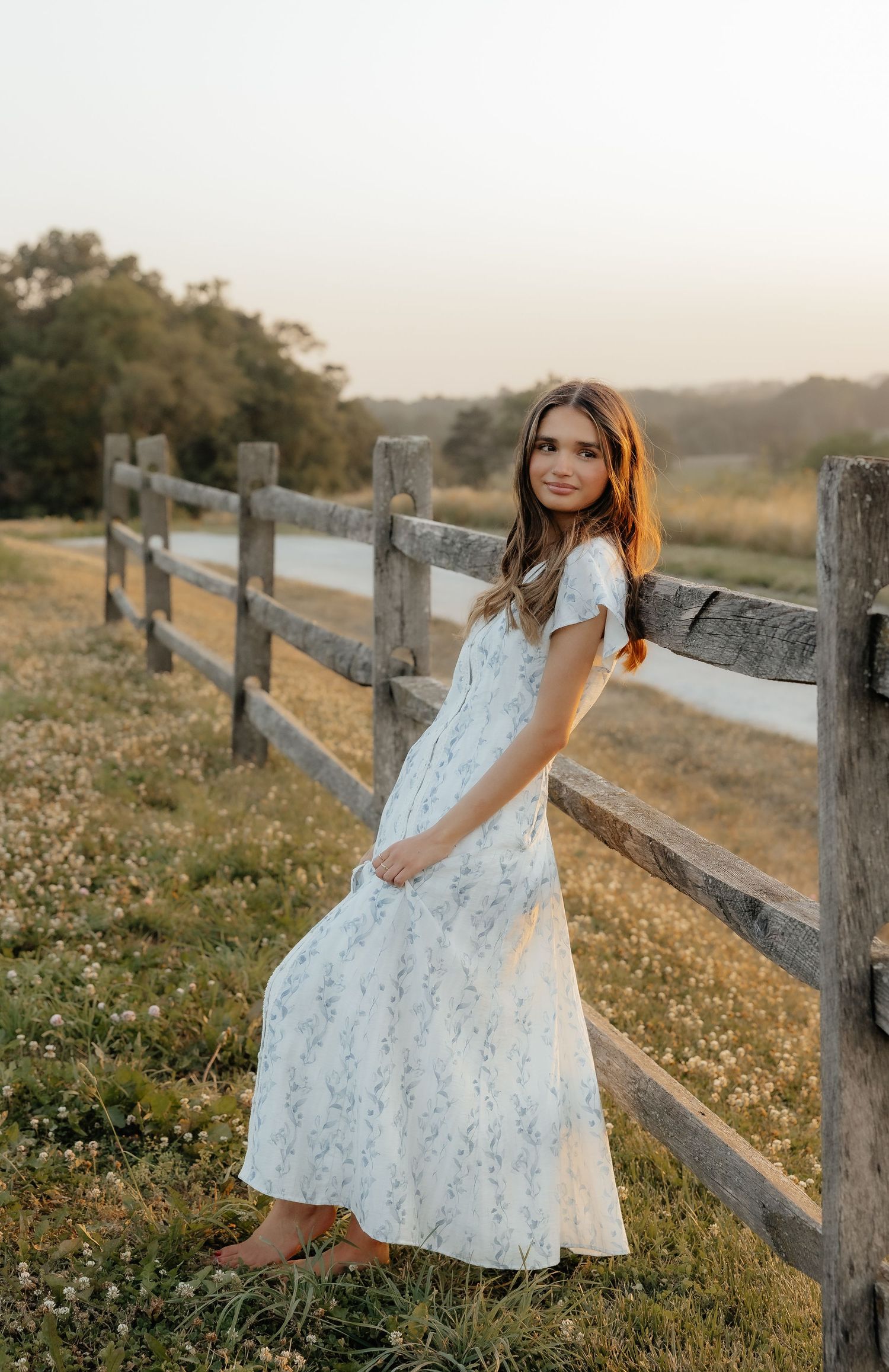 Young woman in white floral maxi dress posing by rustic wooden fence in golden hour sunlight.