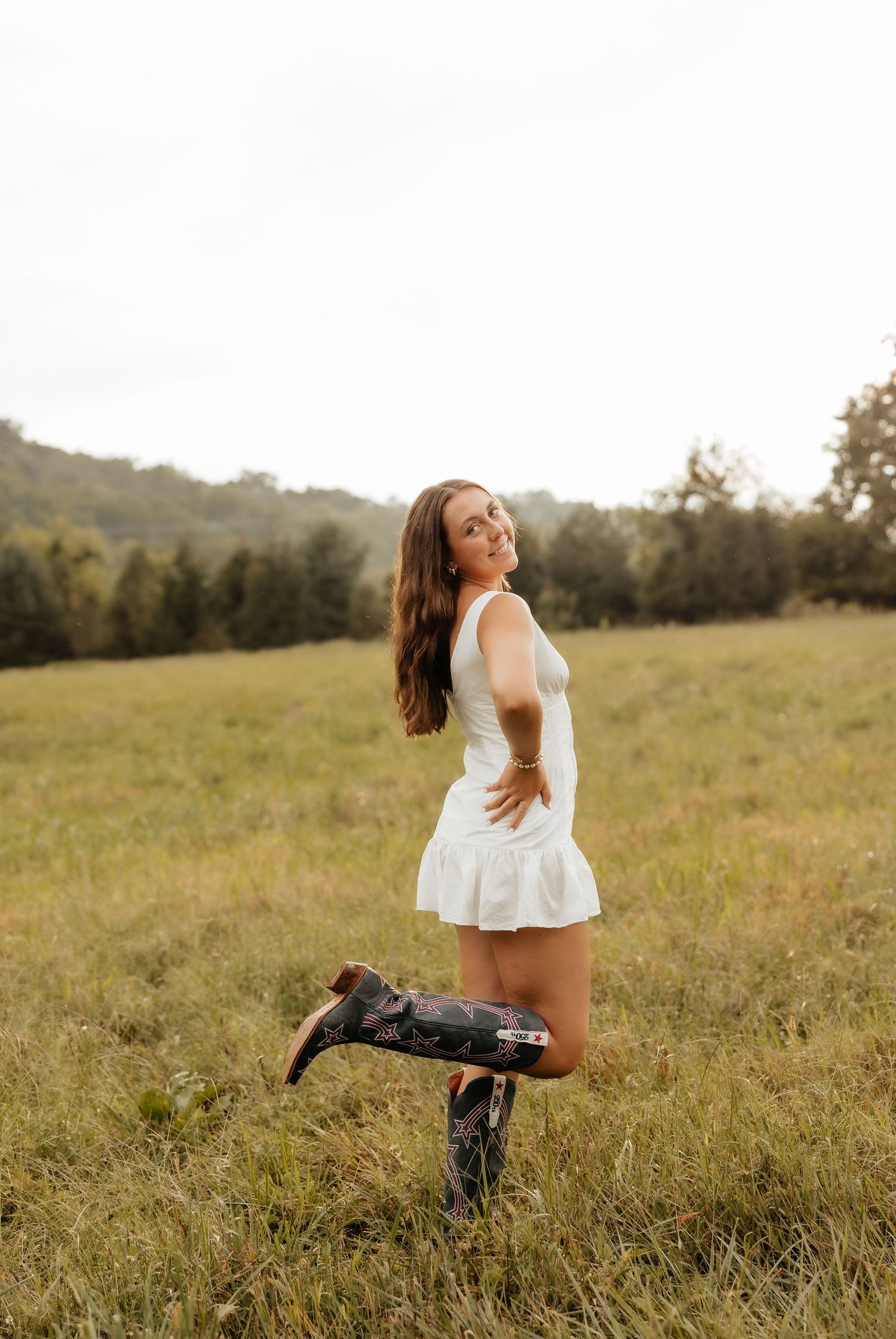 Young woman in white dress and cowboy boots posing in a green field with trees in background.