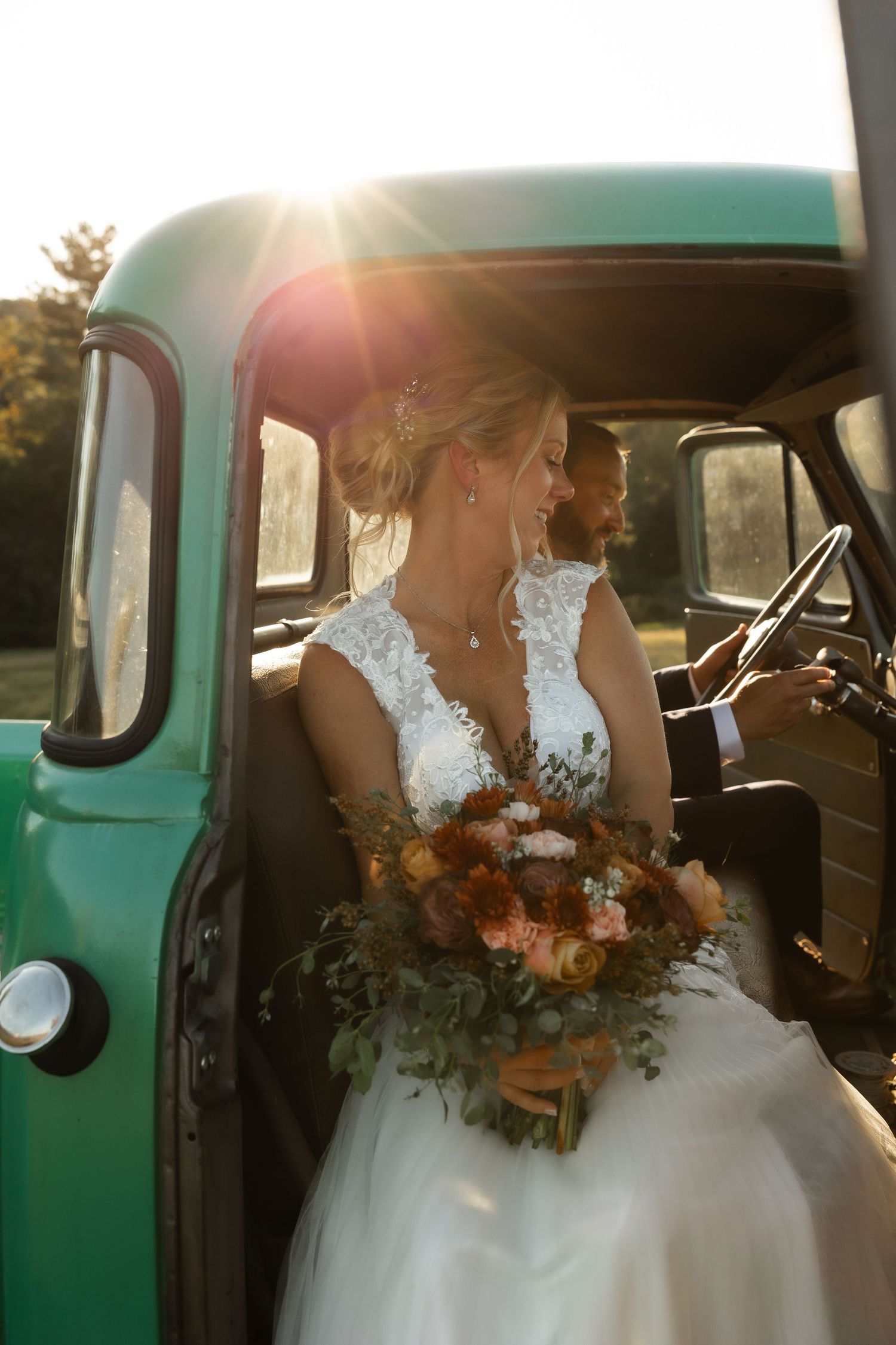 A bride in a white lace dress sits in a vintage turquoise truck holding a fall-colored bouquet at sunset.