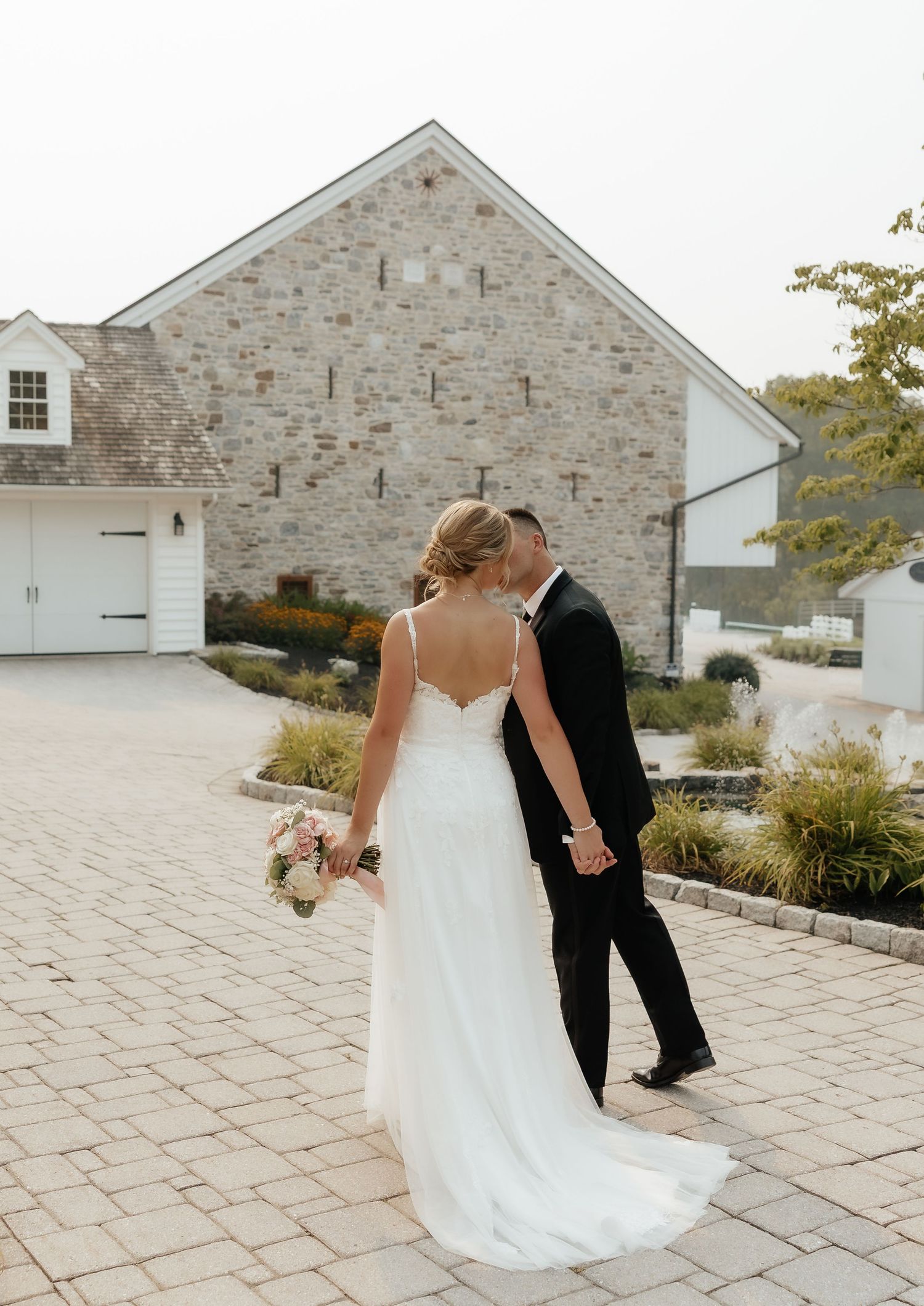 Bride and groom share a kiss outdoors in front of a rustic stone barn venue on their wedding day.