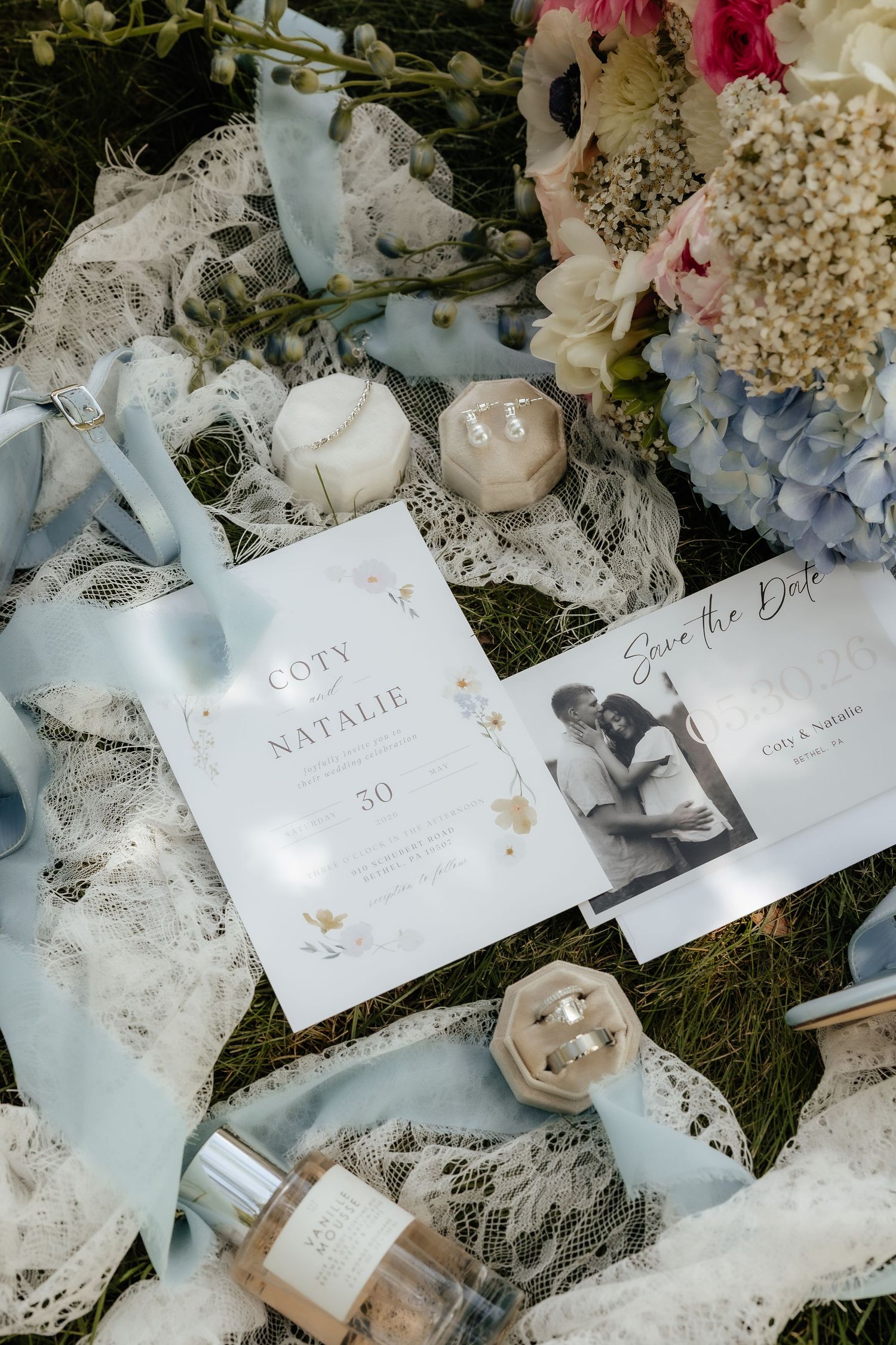 Wedding stationery flat lay with save the date cards, blue ribbons, candles, and floral bouquet on lace fabric.