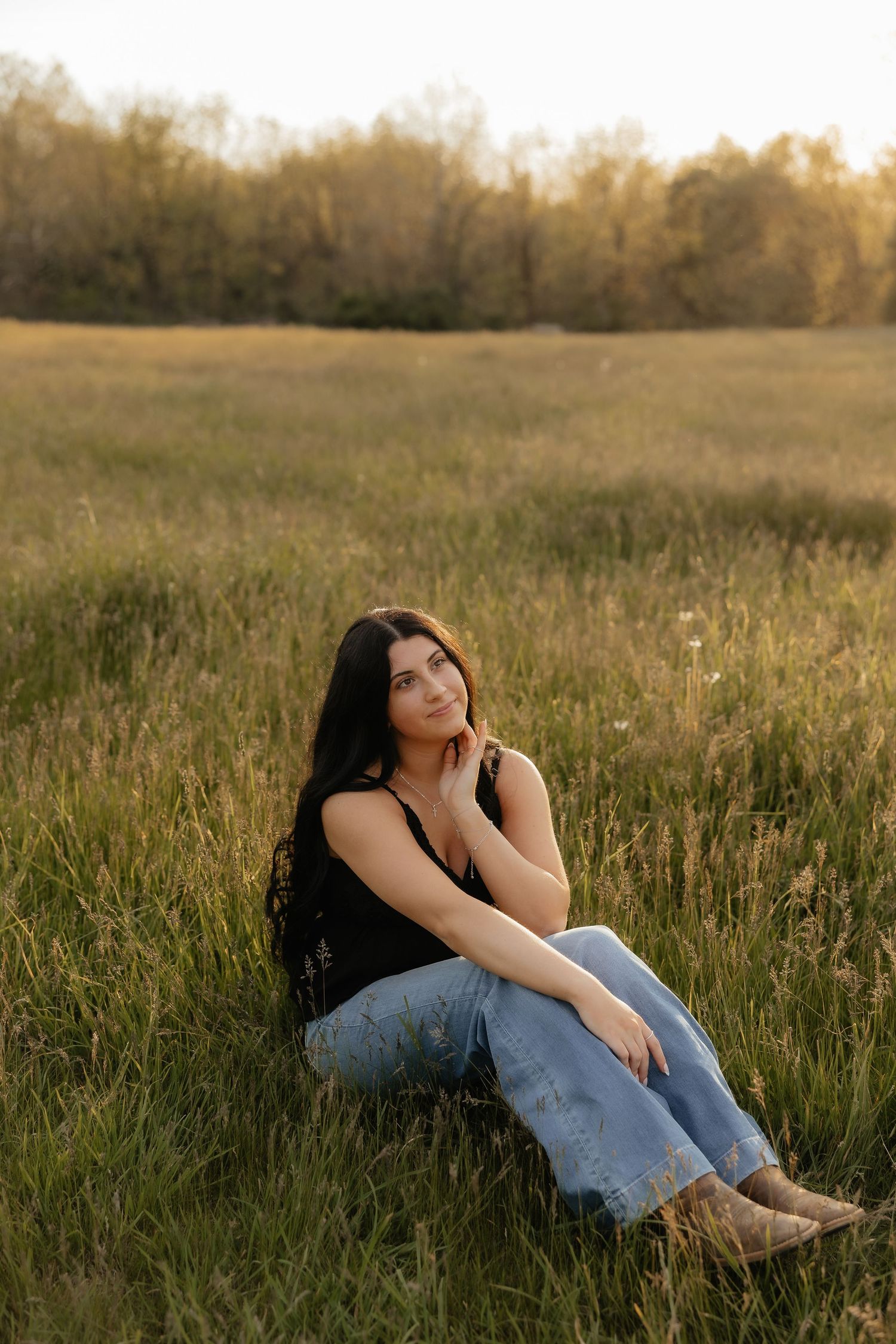Young woman in black tank top and ripped jeans sitting in golden meadow at sunset, gazing upward.