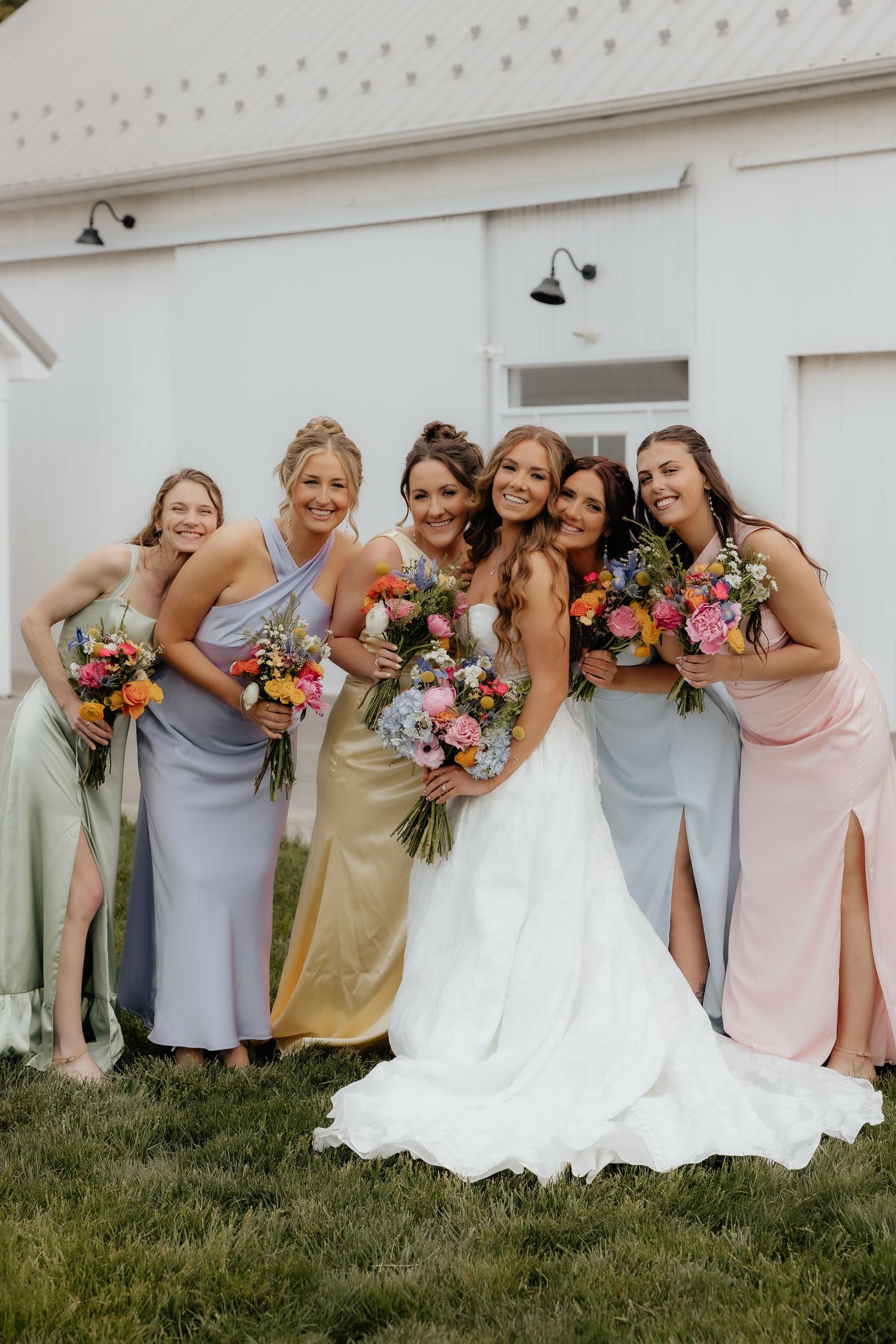 Bride in white gown poses with five bridesmaids in colorful mismatched dresses holding vibrant bouquets outside a white barn.