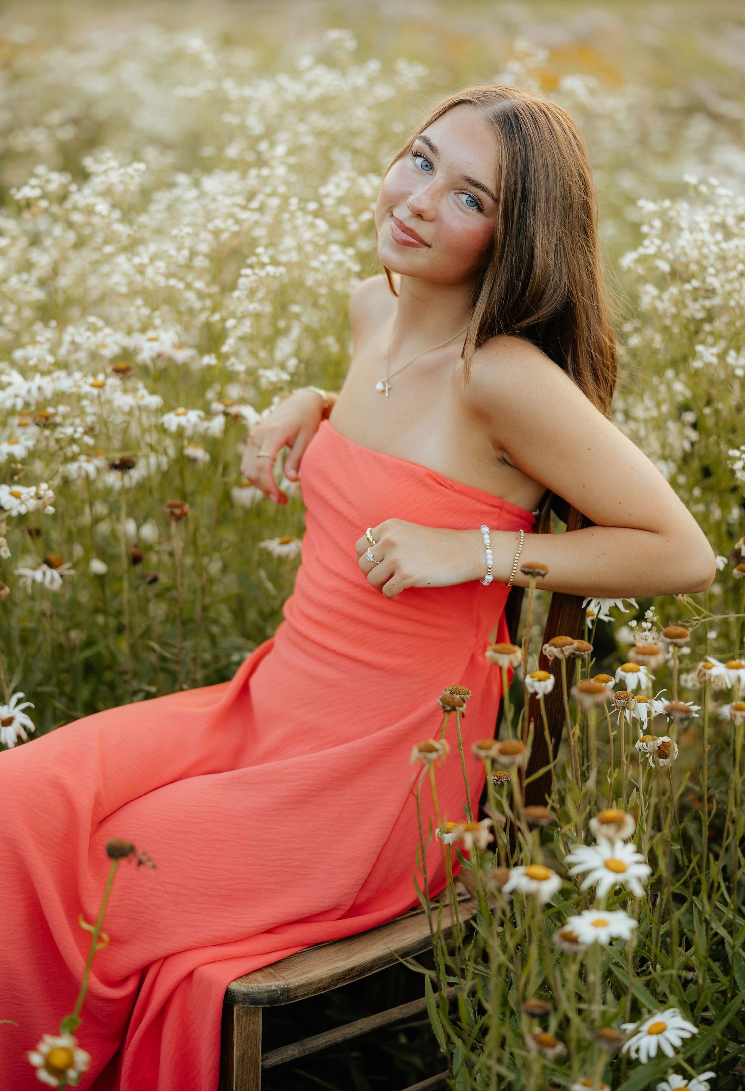 A person in a coral dress poses gracefully on a wooden bench amidst a field of blooming daisies and wildflowers.