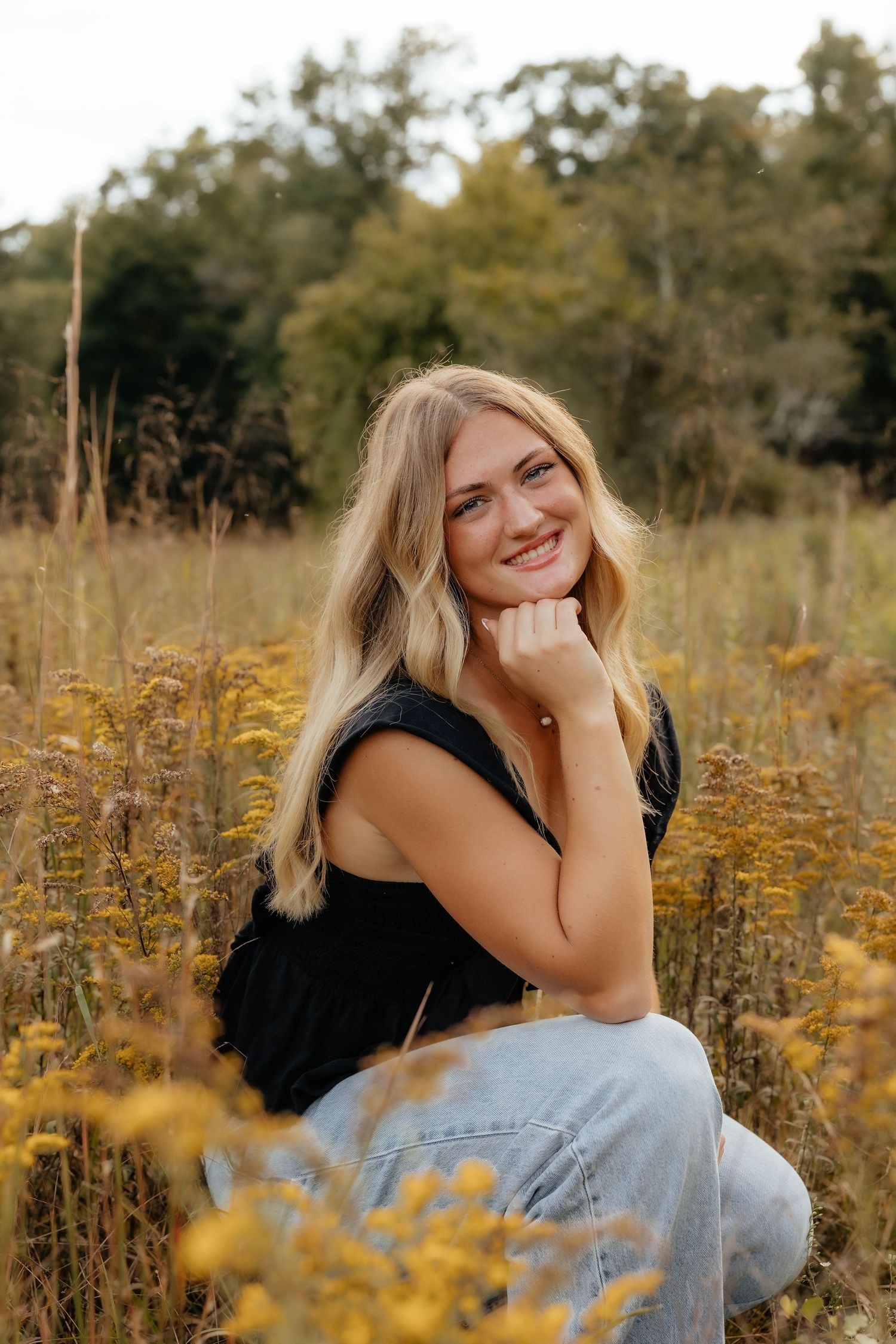 Smiling blonde woman sitting in a golden wildflower field wearing a black top and jeans with trees in the background.