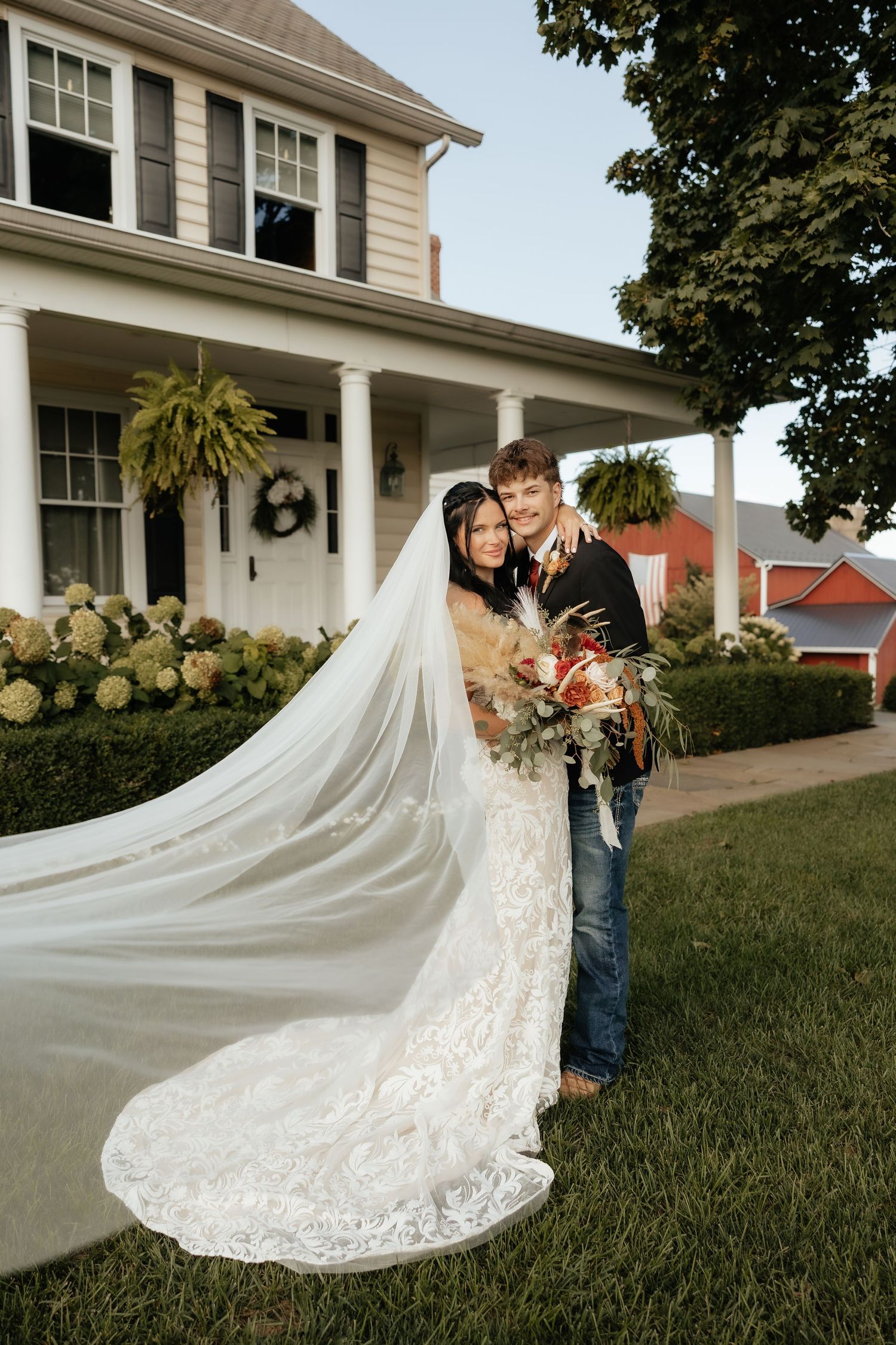 A bride in a lace gown with flowing veil and groom in jeans pose together outside a white farmhouse.
