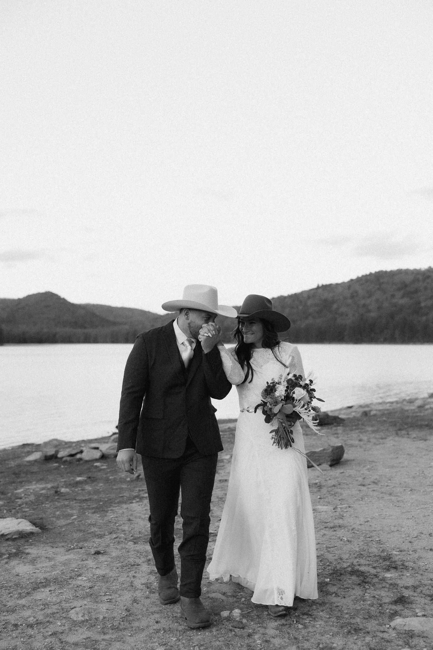 A black and white wedding photo of a couple walking along a lakeside shore.