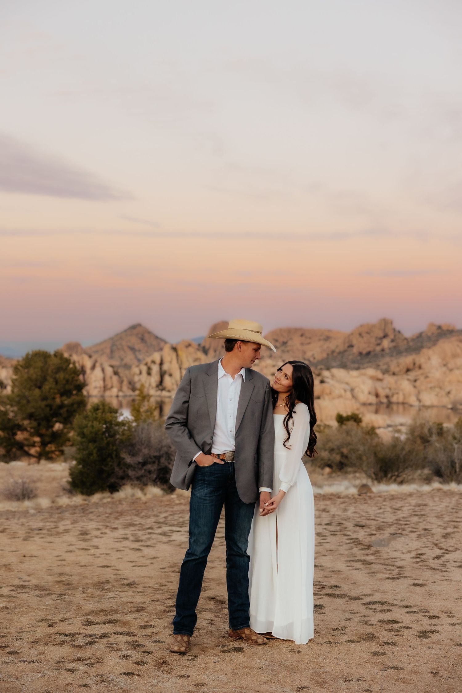 A romantic desert engagement photo with a couple standing together at sunset against rugged mountain backdrop.