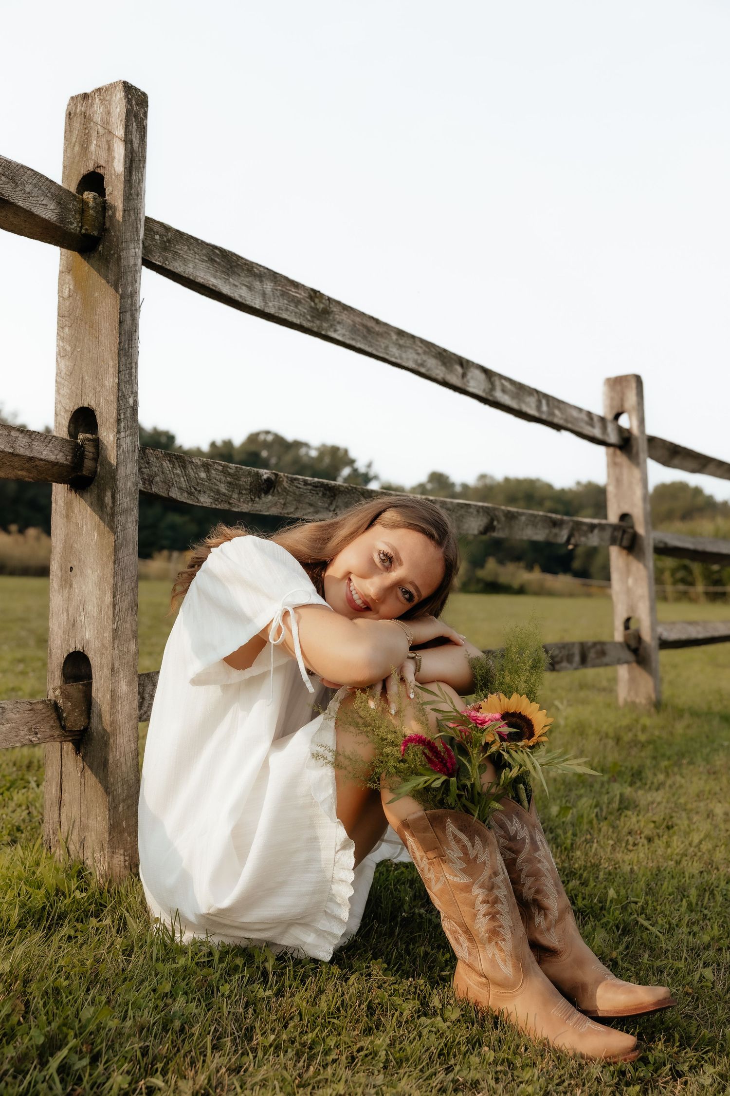 Young woman in white dress and cowboy boots sitting by rustic wooden fence holding sunflowers in a green field.