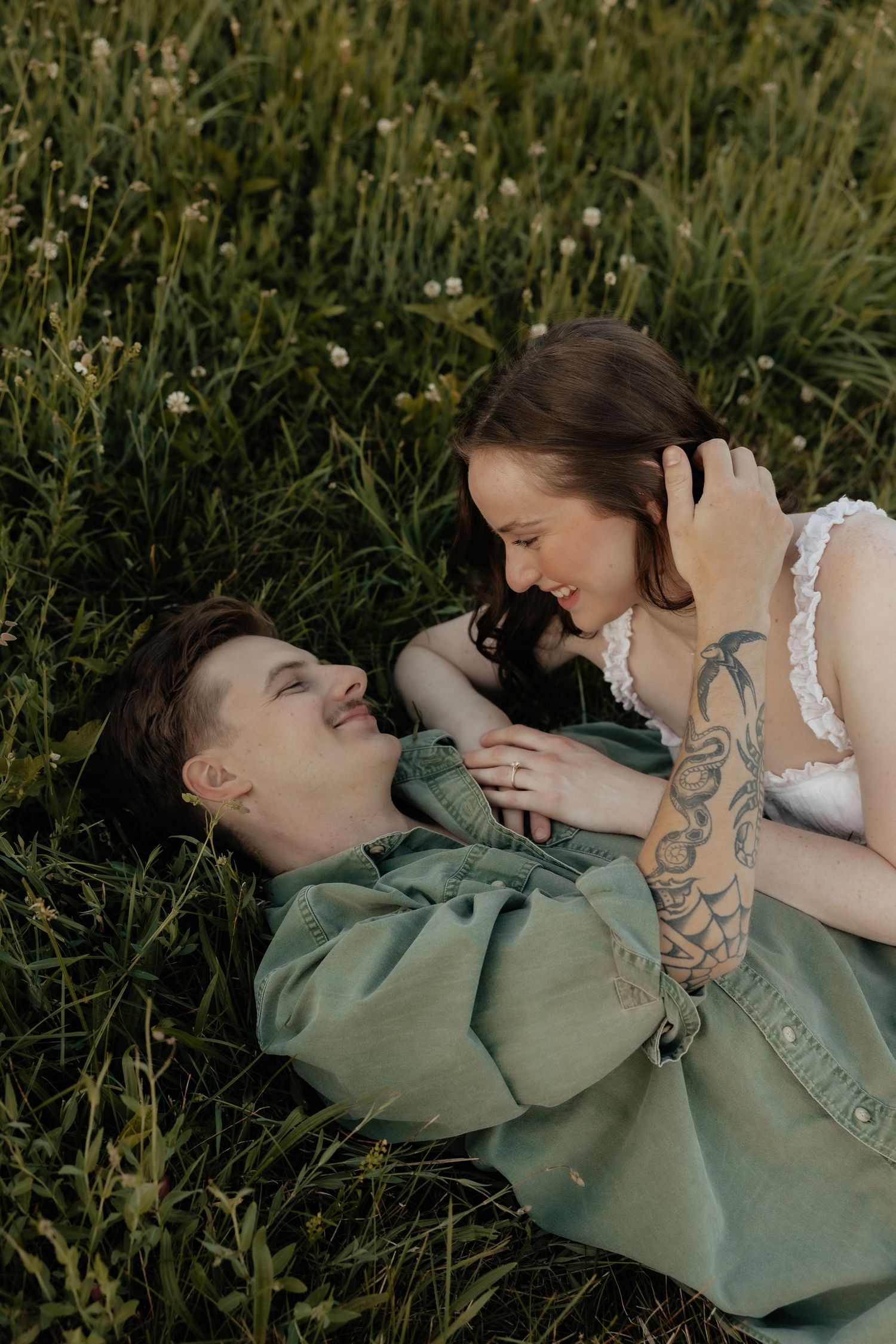 Couple lying in green grass field, smiling at each other, woman in white dress with tattooed arm, man in sage jacket.