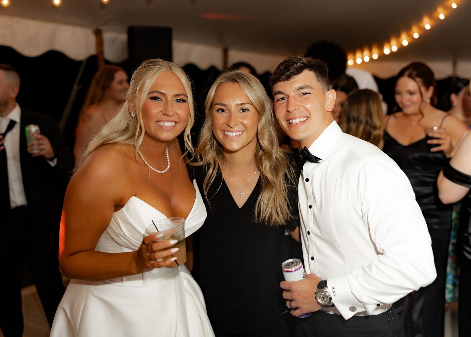 Three well-dressed guests smile and celebrate together at an elegant indoor event with string lights overhead.