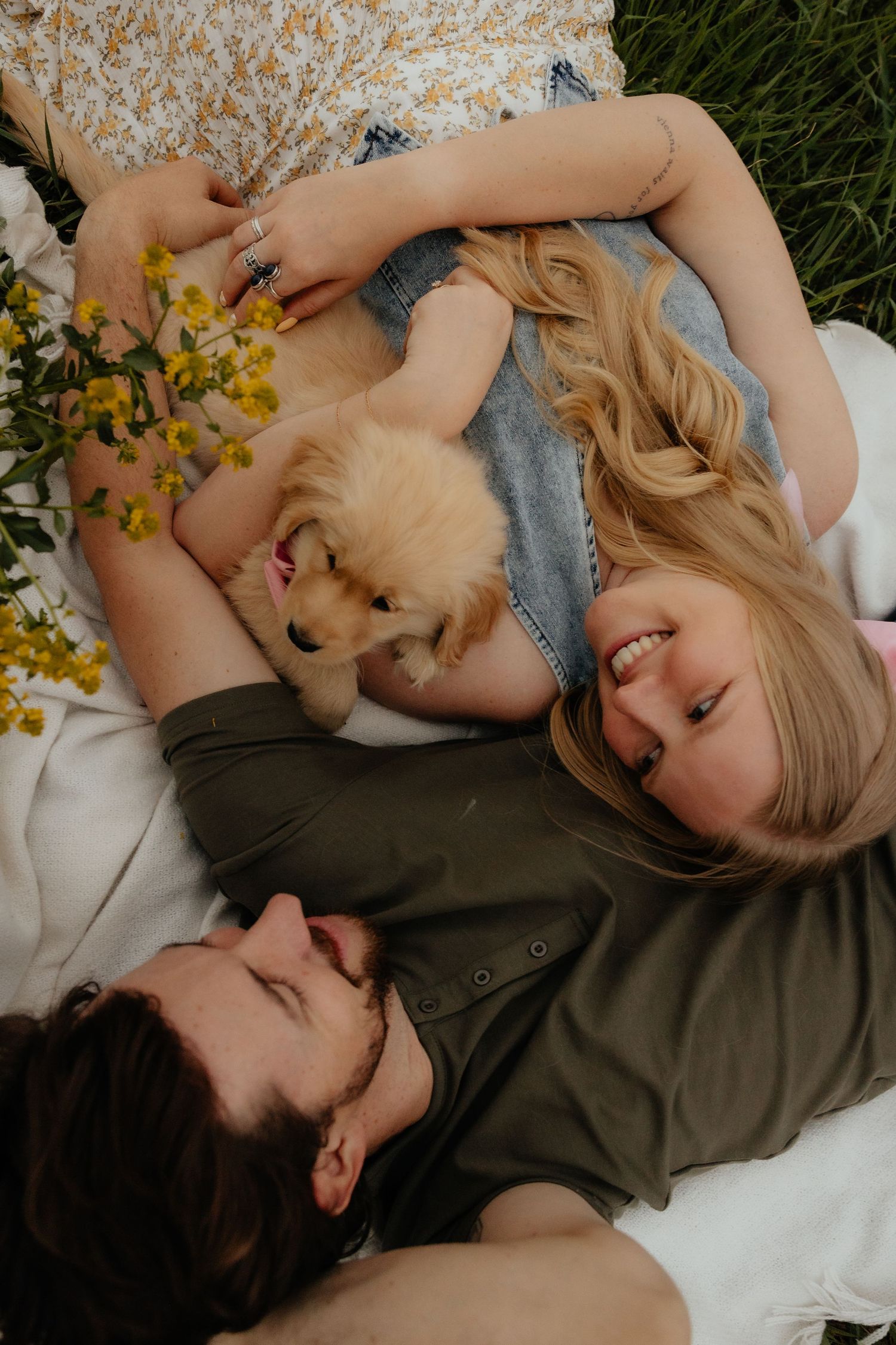 Couple lying on a blanket in grass, smiling and cuddling with an adorable golden retriever puppy outdoors.