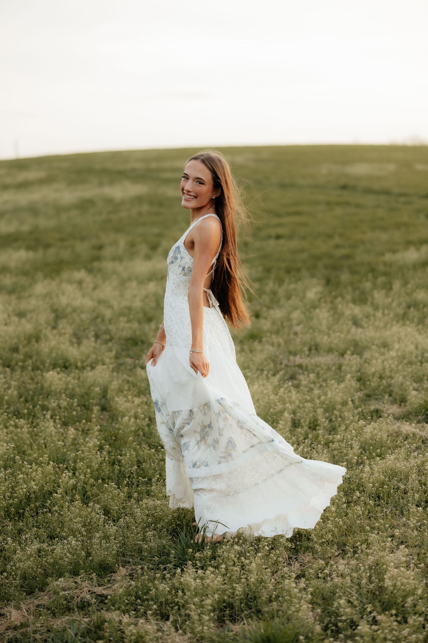 Smiling bride in a flowing white lace gown twirling in a green field during golden hour.