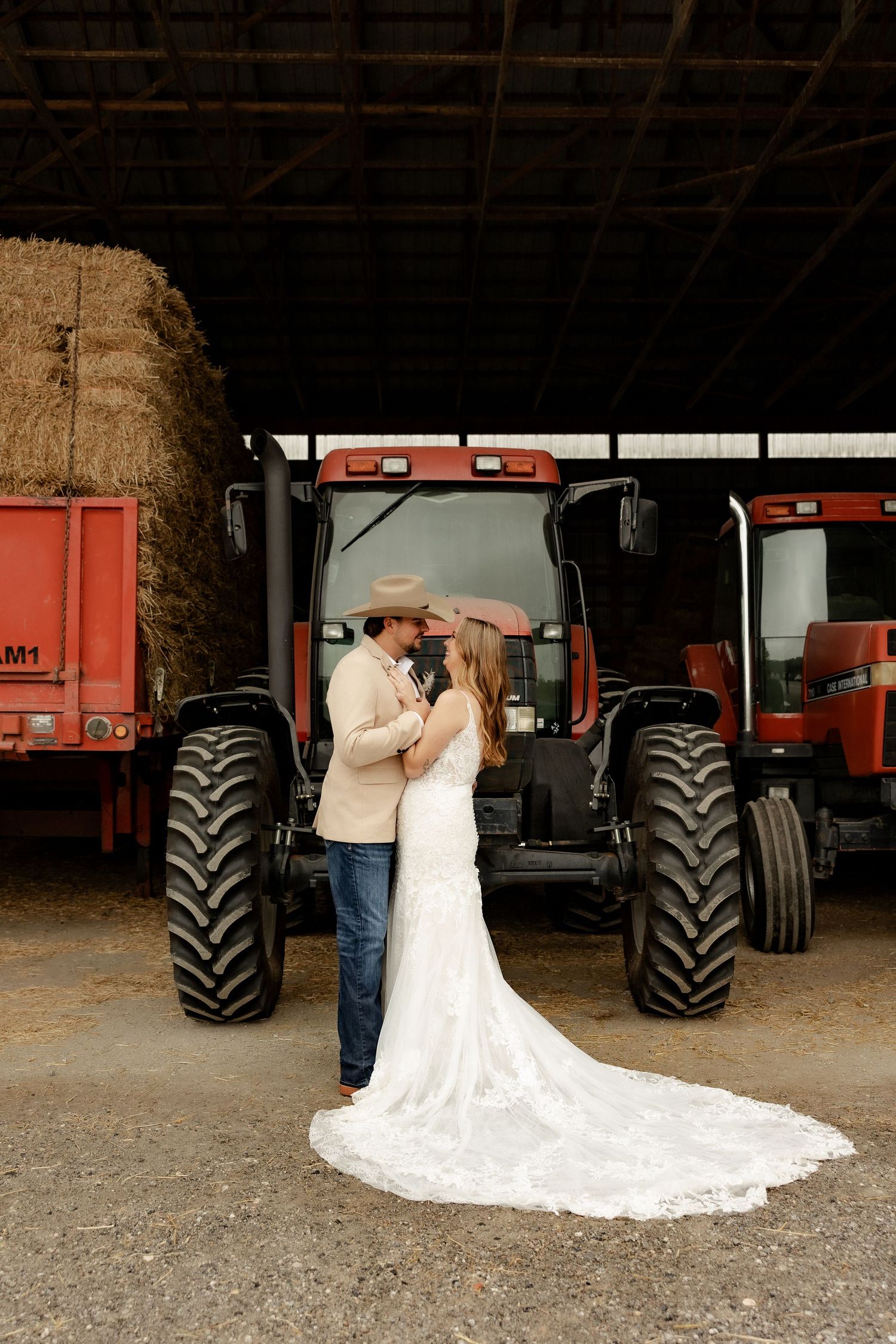 A romantic couple shares an intimate moment in front of red tractors and farm equipment in a rustic barn setting.