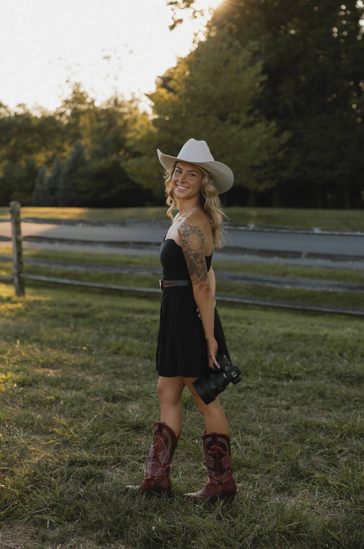 Person in western attire holding camera in grassy field at sunset.