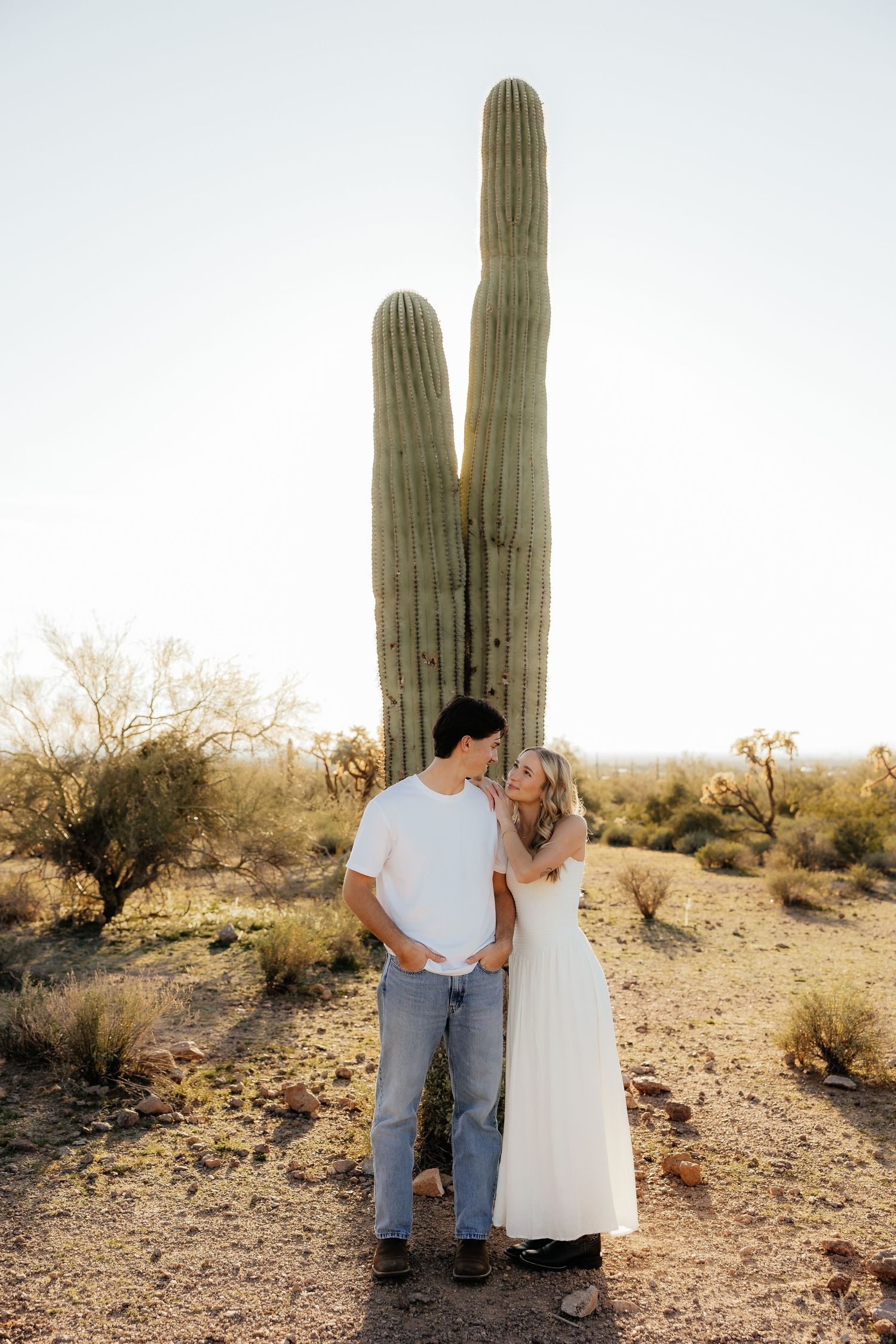 Couple standing together in front of tall saguaro cactus in Arizona desert during golden hour sunset.