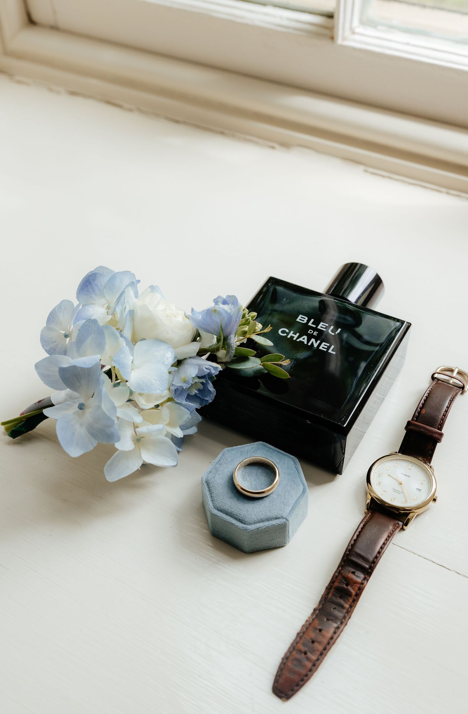 Bleu de Chanel cologne bottle beside blue hydrangeas, a gold ring in velvet box, and brown leather watch on white surface.
