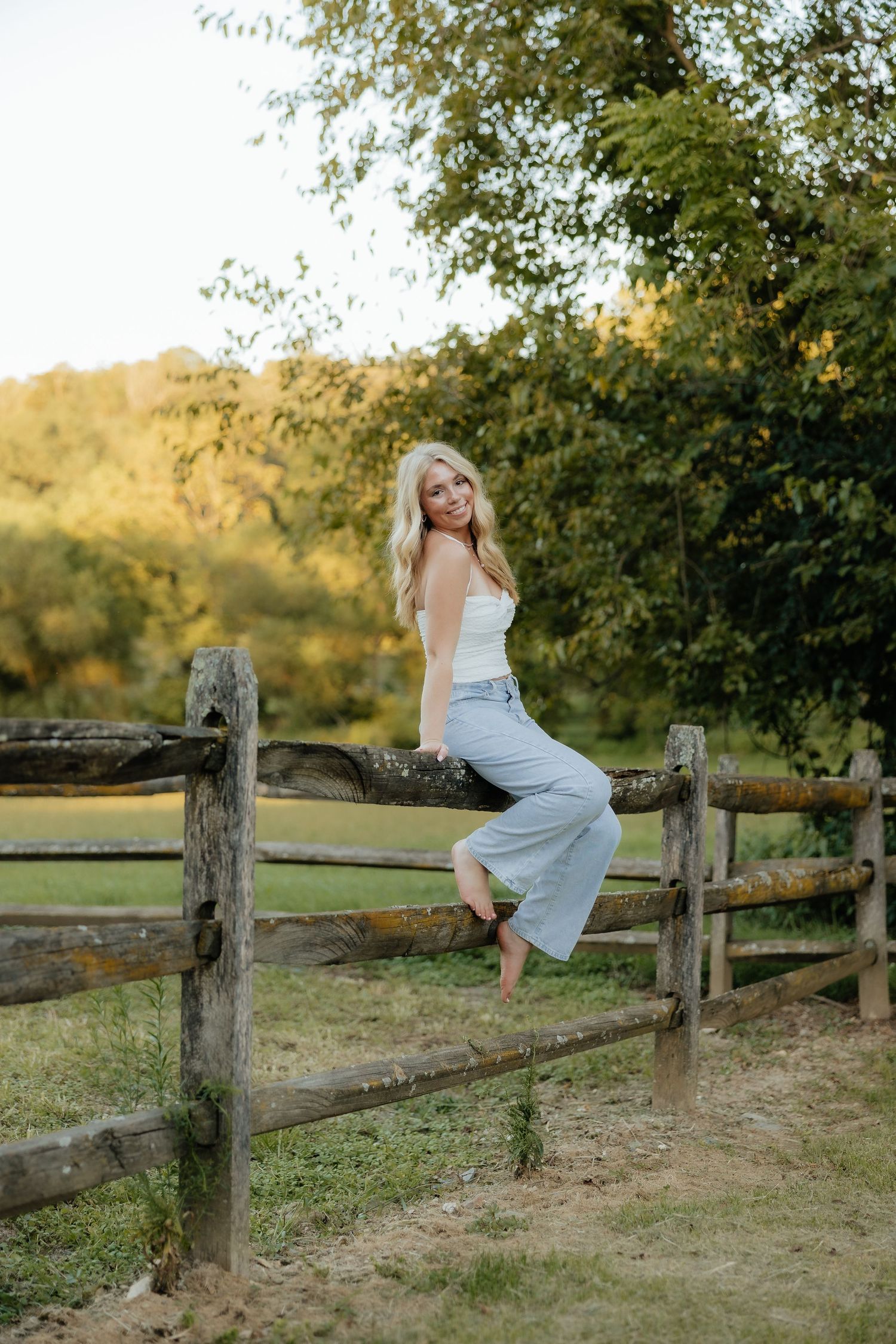 A woman in a flowing white dress sits gracefully on a rustic wooden fence at sunset in a pastoral setting.