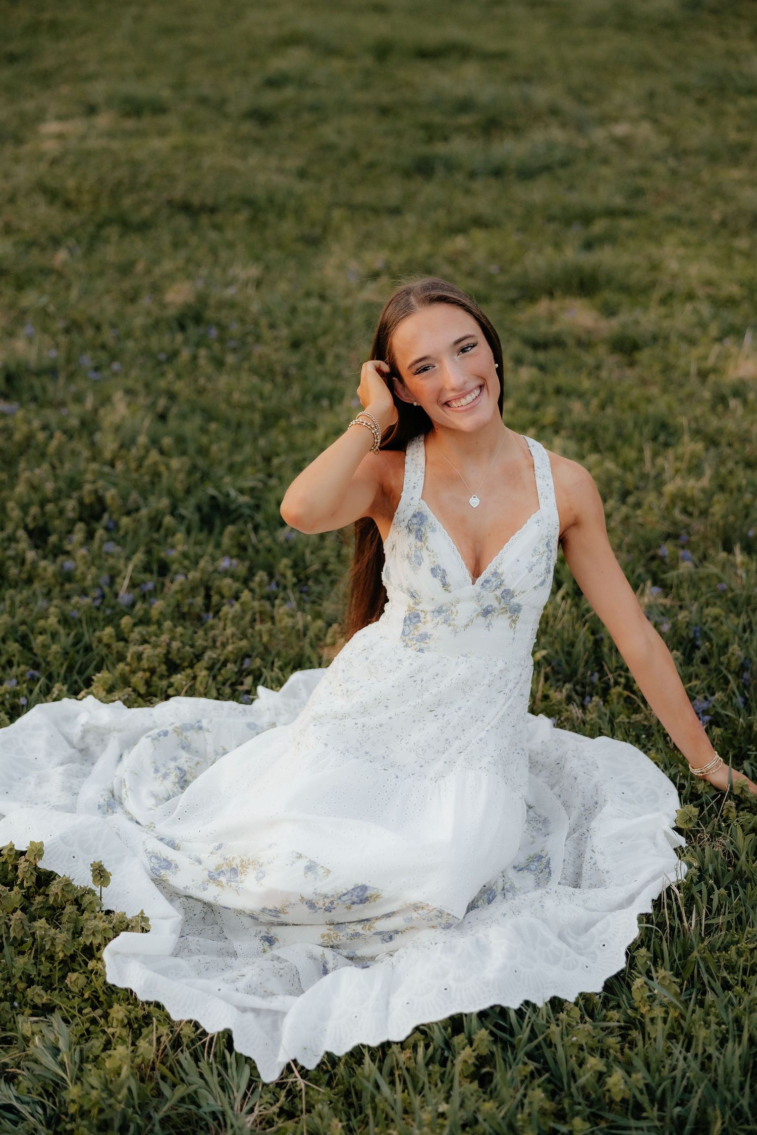 Smiling bride in white floral gown sitting gracefully on green grass field in golden hour light.