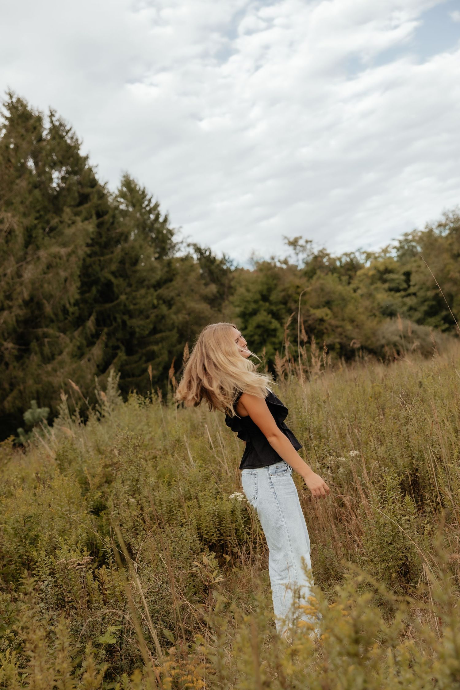 Blonde woman in black top and jeans tossing her hair in a lush green field with trees in the background.