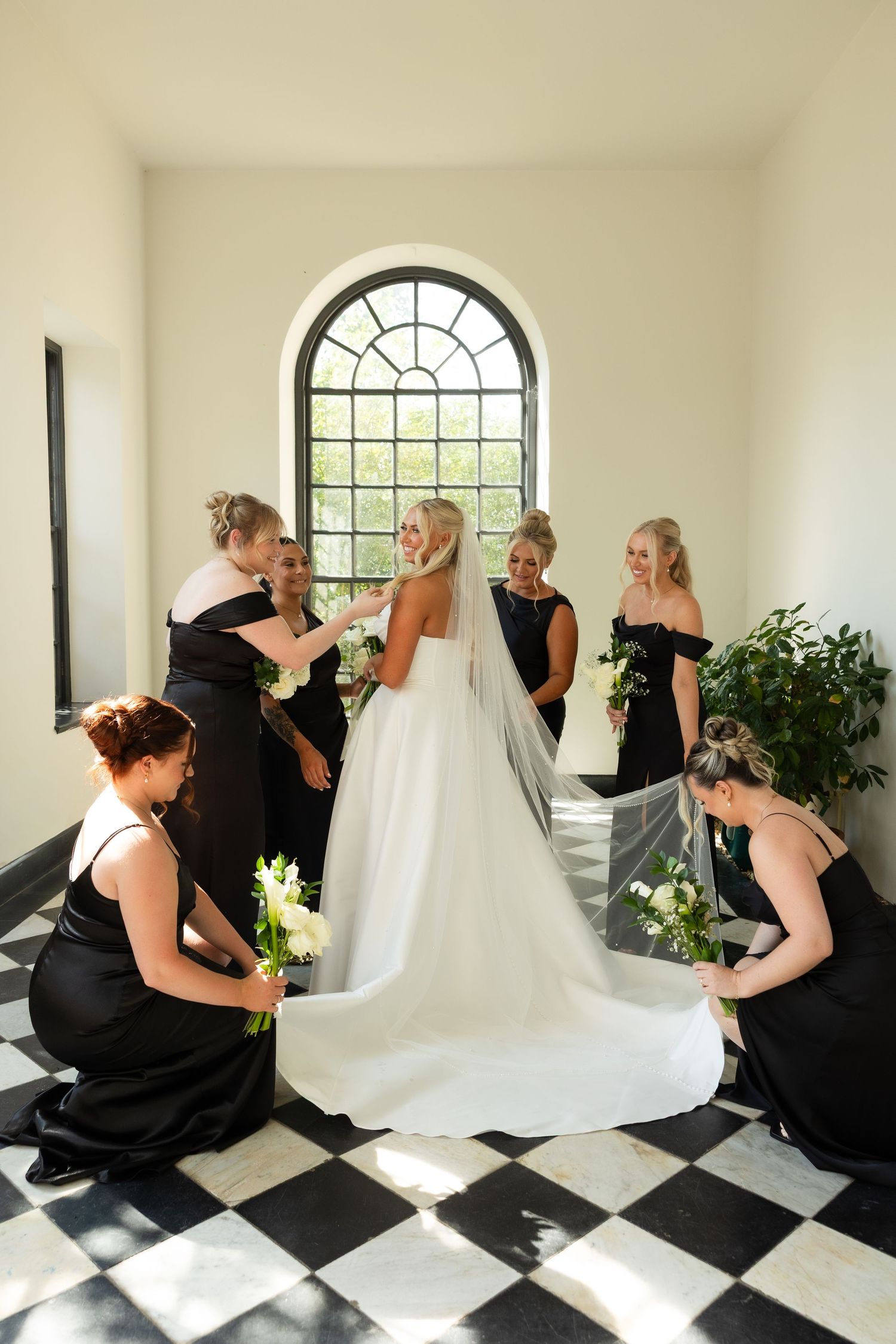 A wedding party helps adjust a bride's long white train on a black and white checkered floor beneath an arched window.