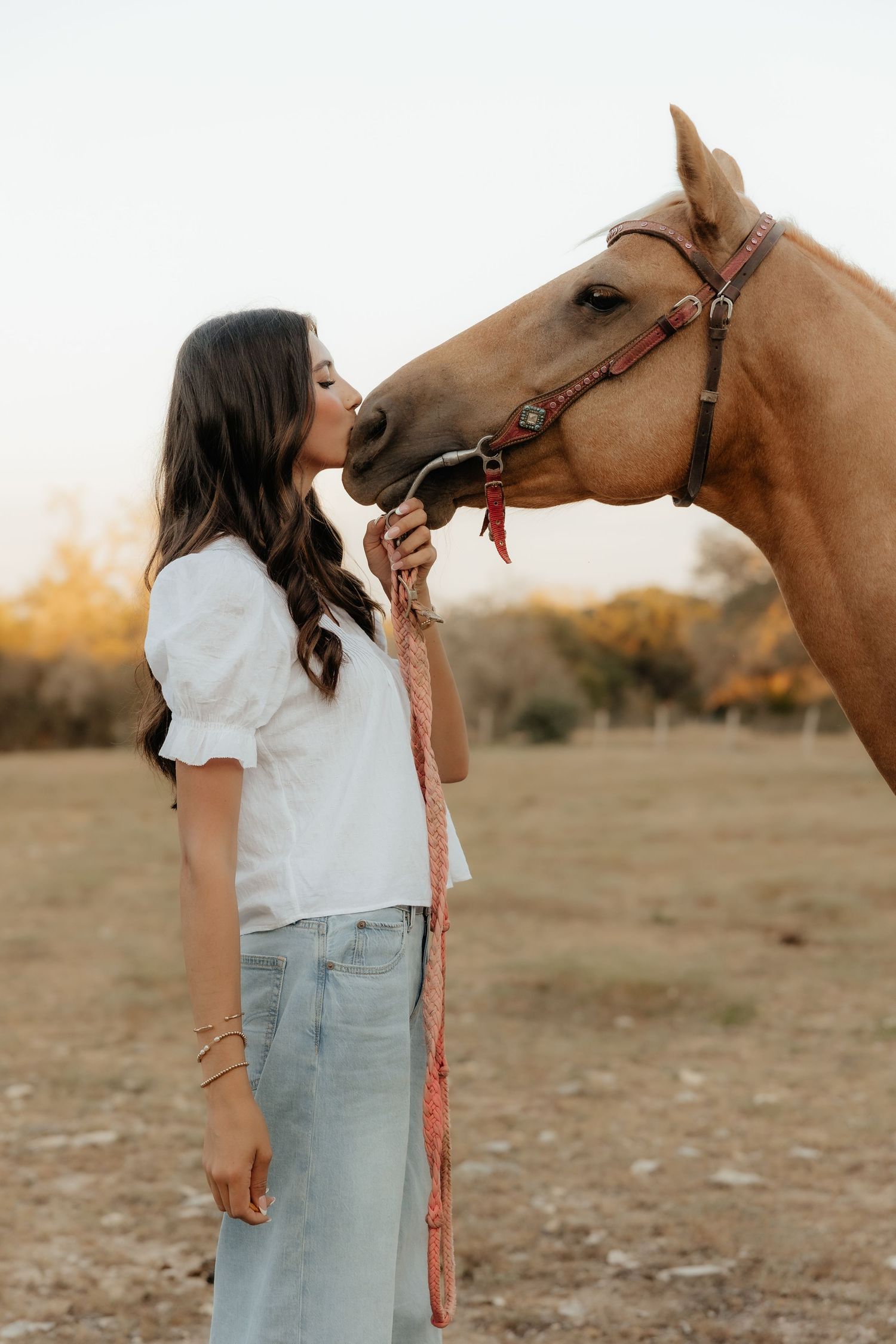 A young woman in a white top and jeans shares a tender kiss with a palomino horse in a sunlit field.