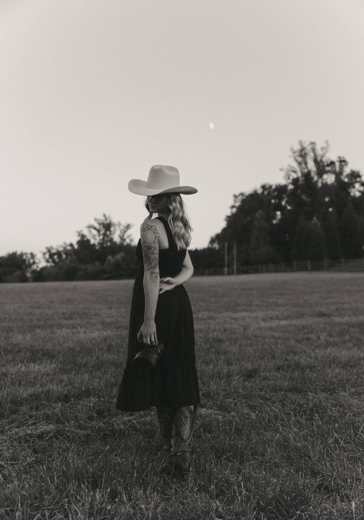 Black and white photo of a woman in cowboy hat and dark dress standing in a field at dusk.