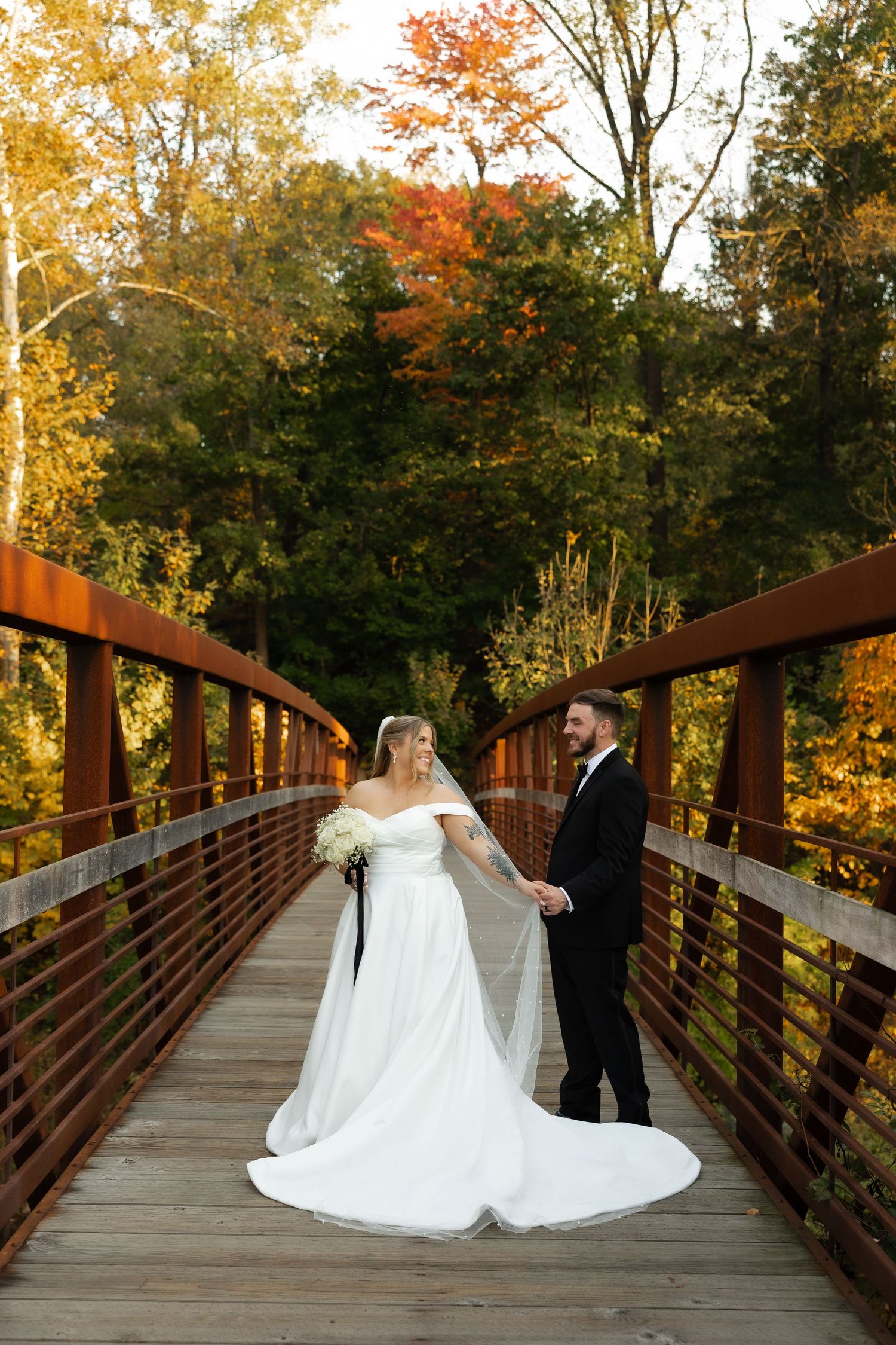 A couple in wedding attire share a romantic moment on a wooden bridge surrounded by autumn foliage at sunset.