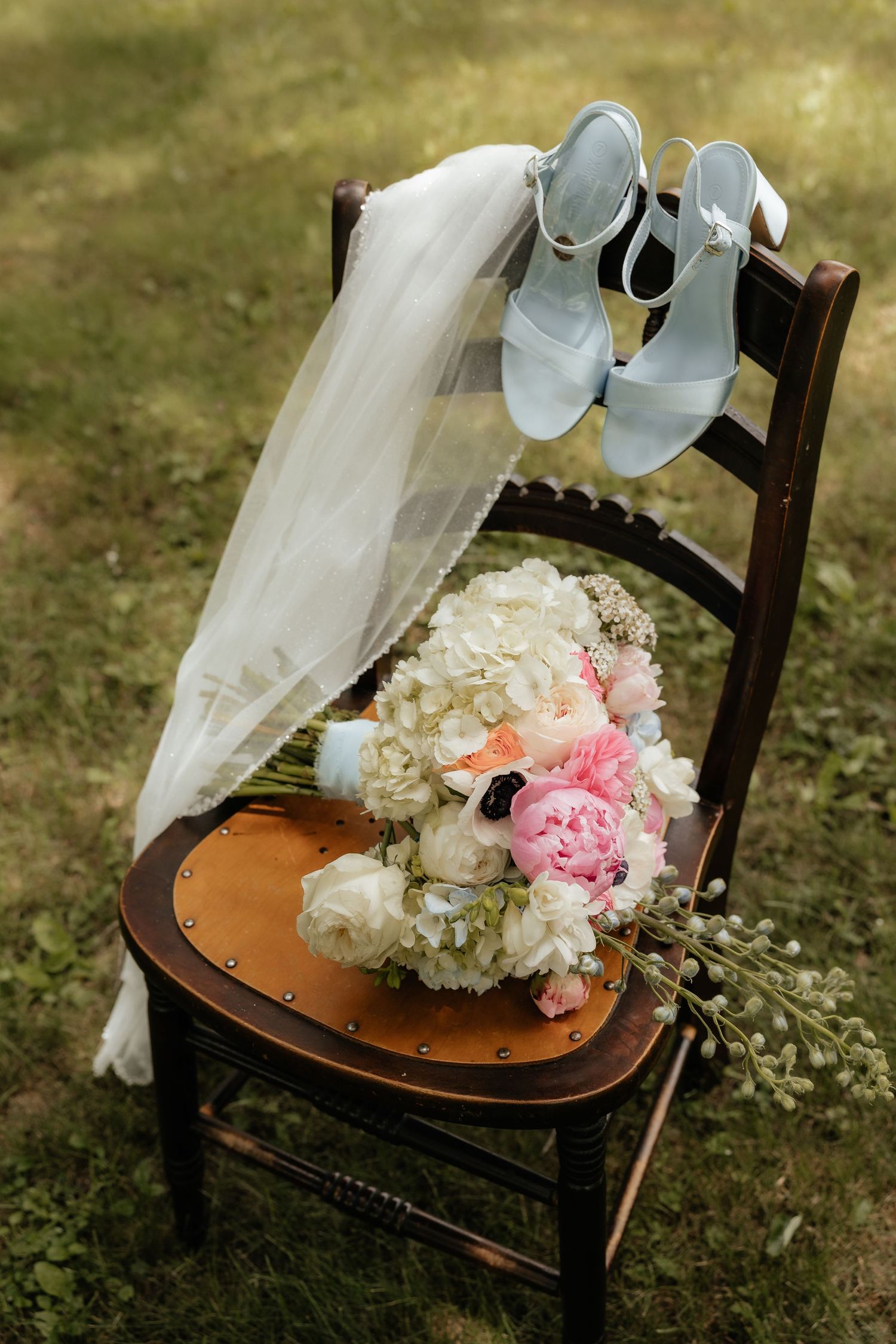 Bridal bouquet of pink peonies and white hydrangeas on wooden chair with blue heels and flowing veil outdoors.