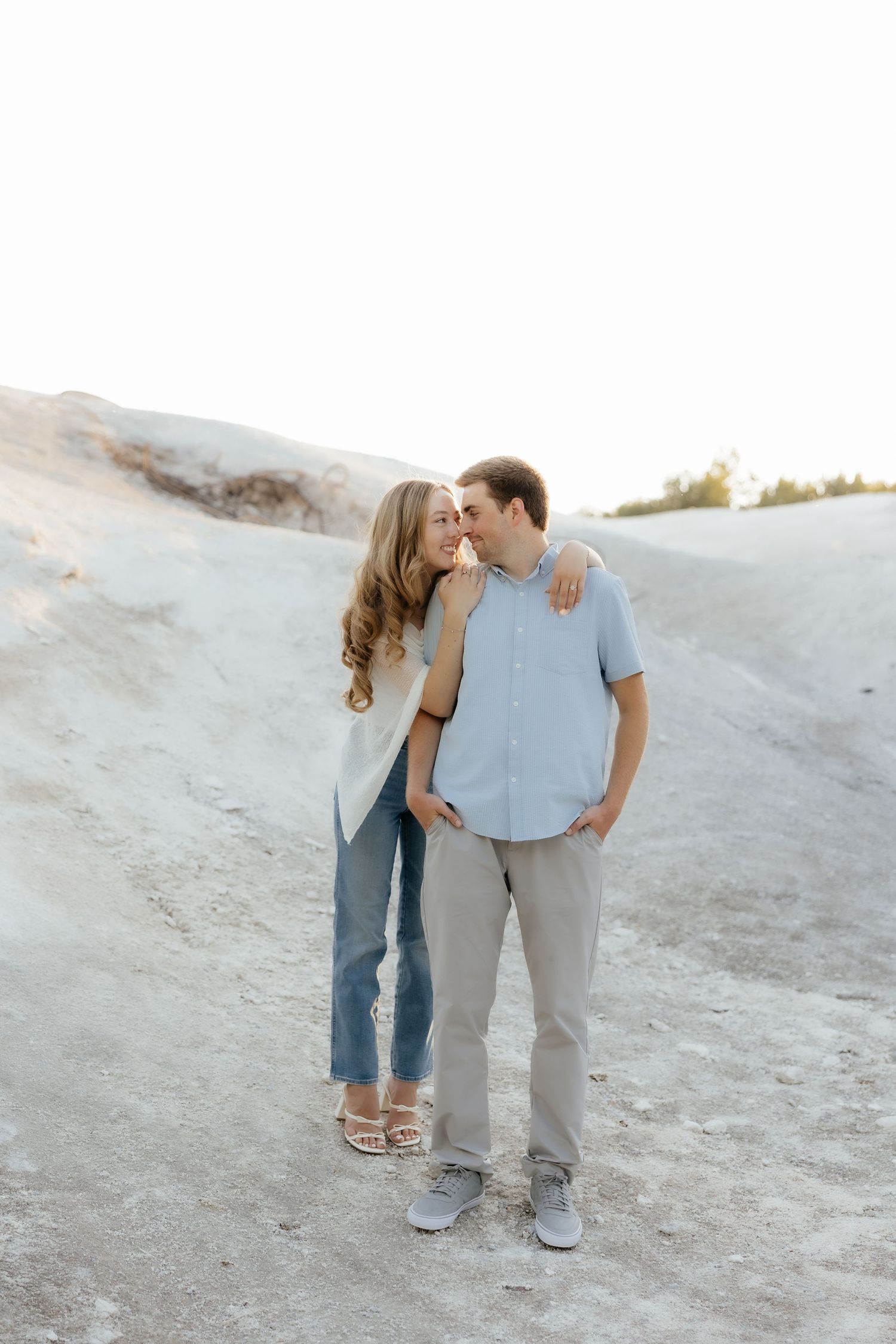 A couple embraces and smiles on a sandy hillside, the woman leaning into the man in casual blue and neutral tones.