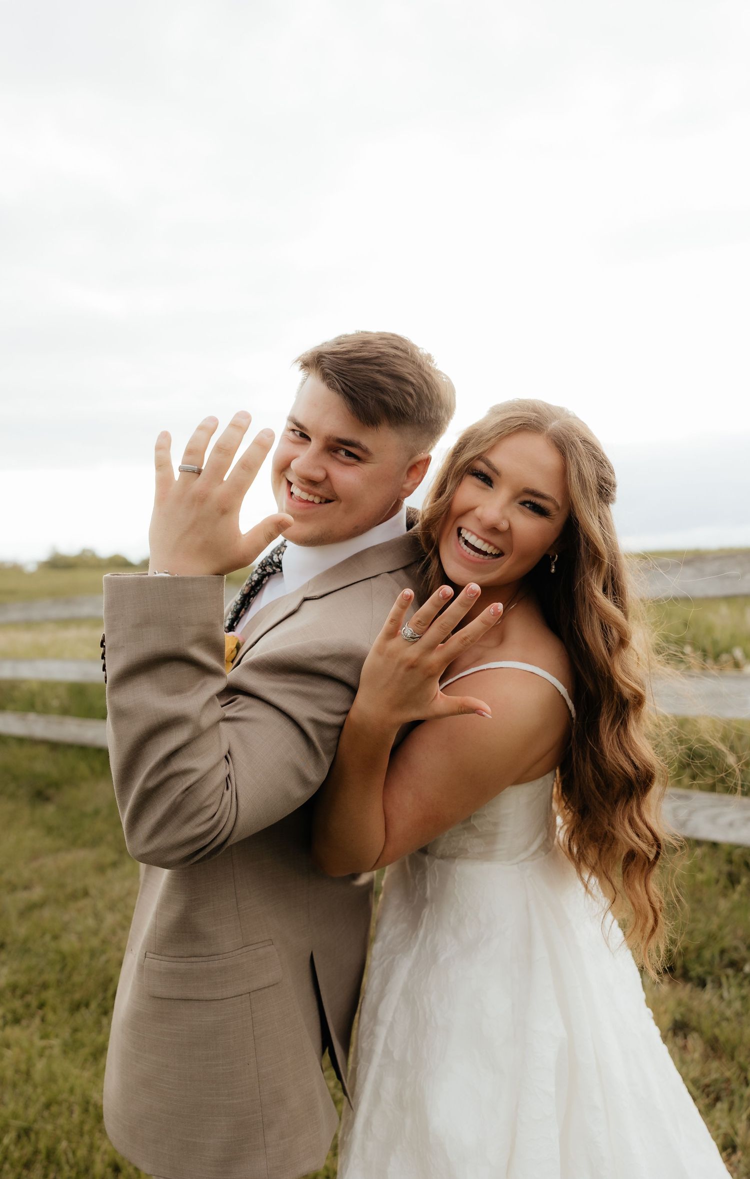 Smiling newlyweds in wedding attire show off their rings outdoors near a wooden fence on their wedding day.