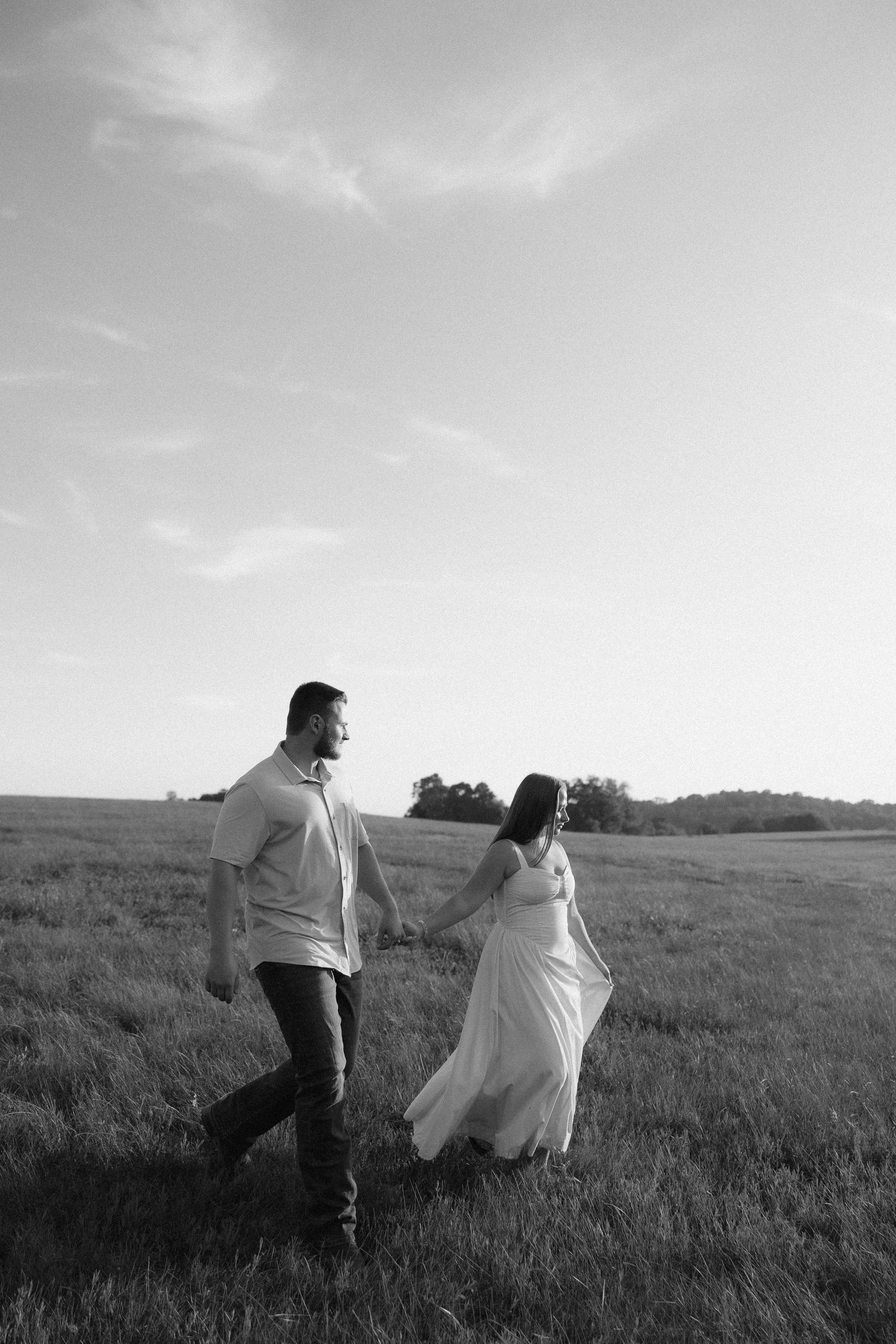 Black and white photo of a couple holding hands walking through an open field, woman in flowing dress, trees in background.