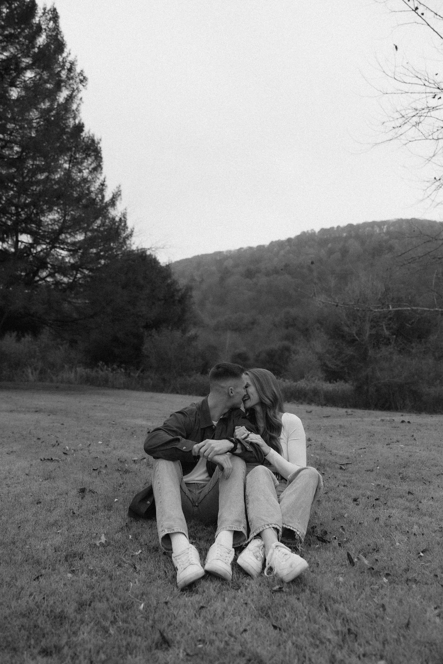 A couple lies together in a grassy field with mountains and trees visible in the background.