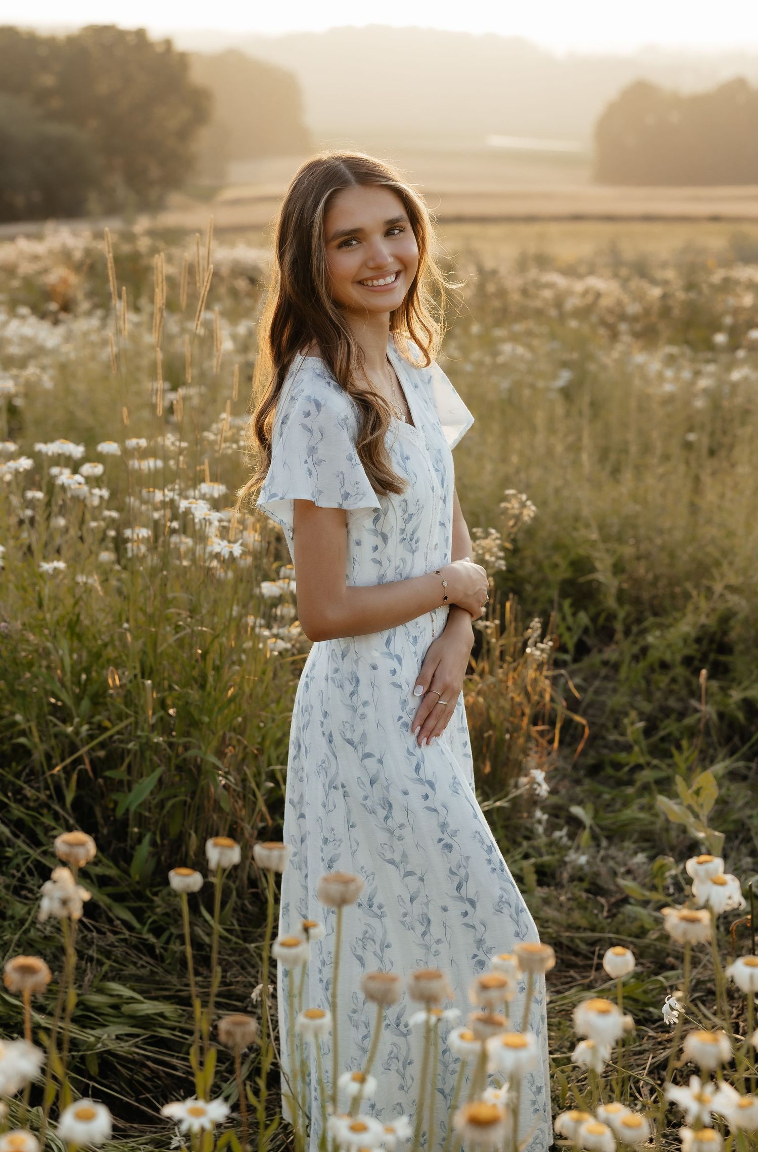 Young woman in a floral maxi dress smiling in a wildflower meadow during golden hour sunset.